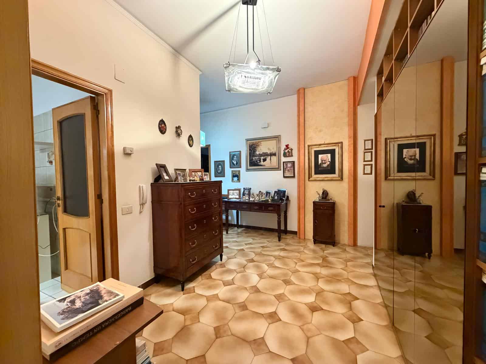 Interno di un soggiorno elegante con mobili in legno, pareti decorate e pavimento in piastrelle esagonali, illuminato da lampadario centrale.
