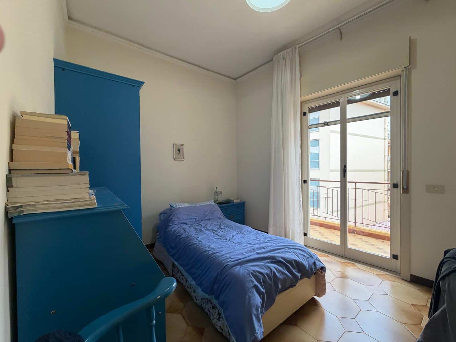 Letto singolo con coperta blu in camera luminosa con balcone e tende bianche, ideale per affitti brevi o soggiorni di relax.