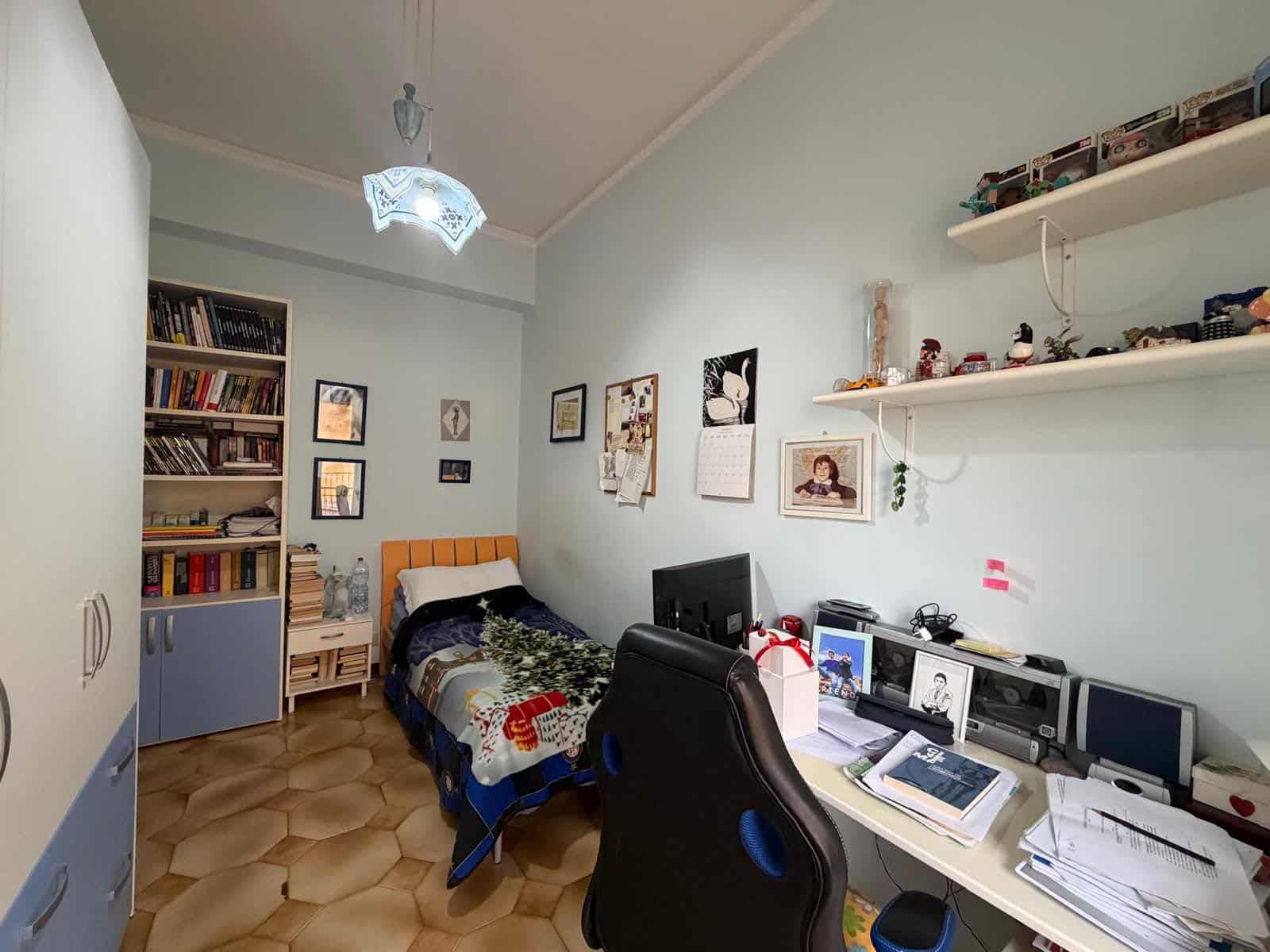 Letto singolo con coperta colorata, scrivania con computer e libri, pareti decorate con quadri e mensole con oggetti decorativi, in una stanza da letto luminosa e accogliente.