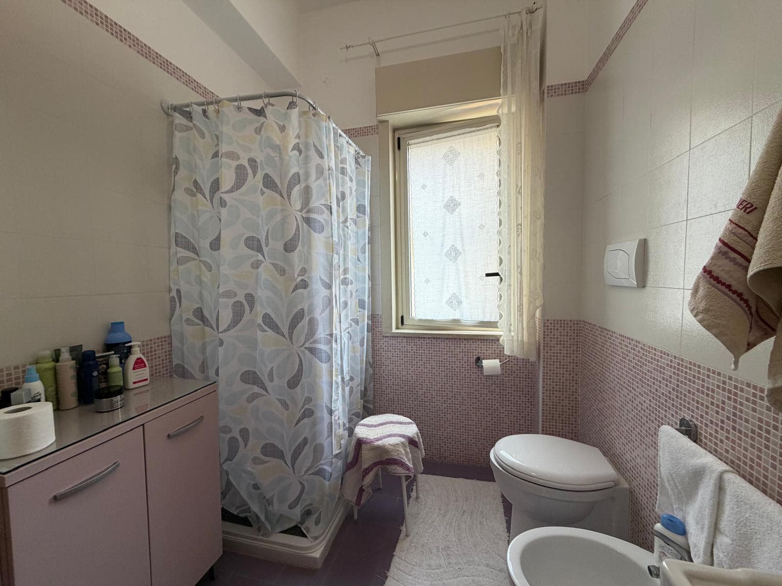 Doccia in bagno con tenda floreale, finestra luminosa e accessori per la cura personale, ambiente pulito e funzionale.