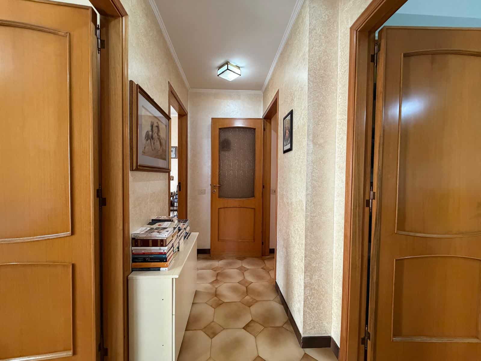 Ampio corridoio in stile classico con pareti beige, pavimento in piastrelle esagonali e porte in legno massello, illuminato da plafoniera moderna.