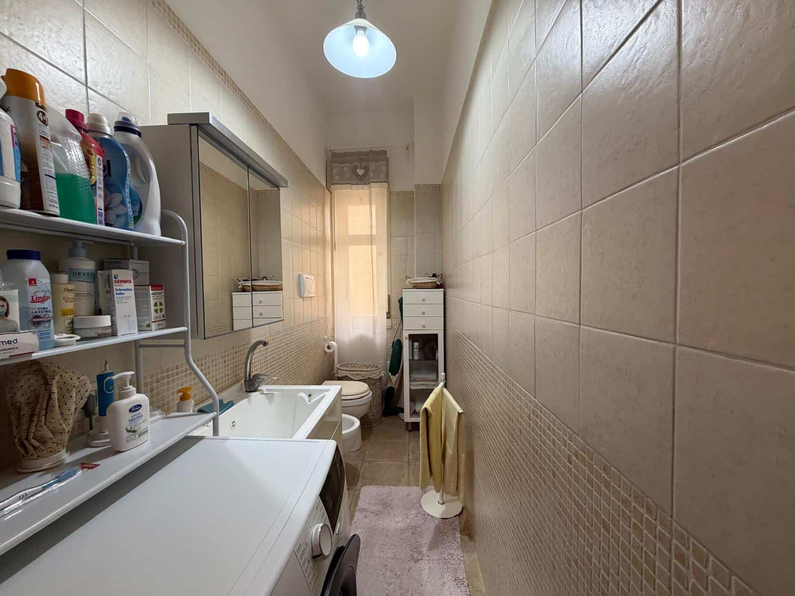 Lavanderia spaziosa con lavatrice, scaffali con detersivi e asciugamani, illuminata da luce naturale e lampada a soffitto, ideale per il lavaggio e la cura dei tessuti domestici.