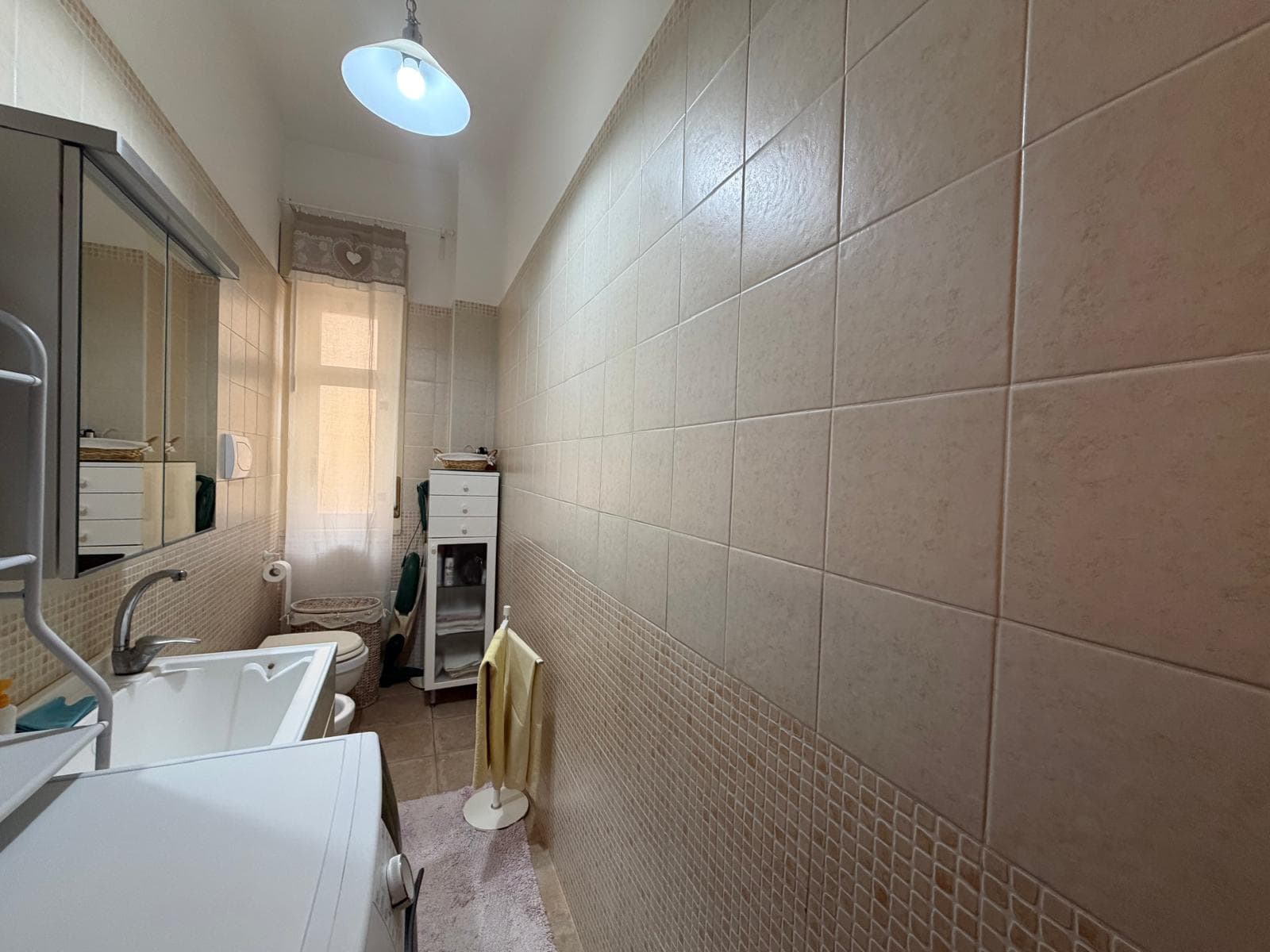 Lavatrice e asciugatrice in un'area lavanderia luminosa e funzionale, con pareti rivestite di piastrelle beige e finestra che permette l'ingresso di luce naturale, ideale per la cura dei vestiti.