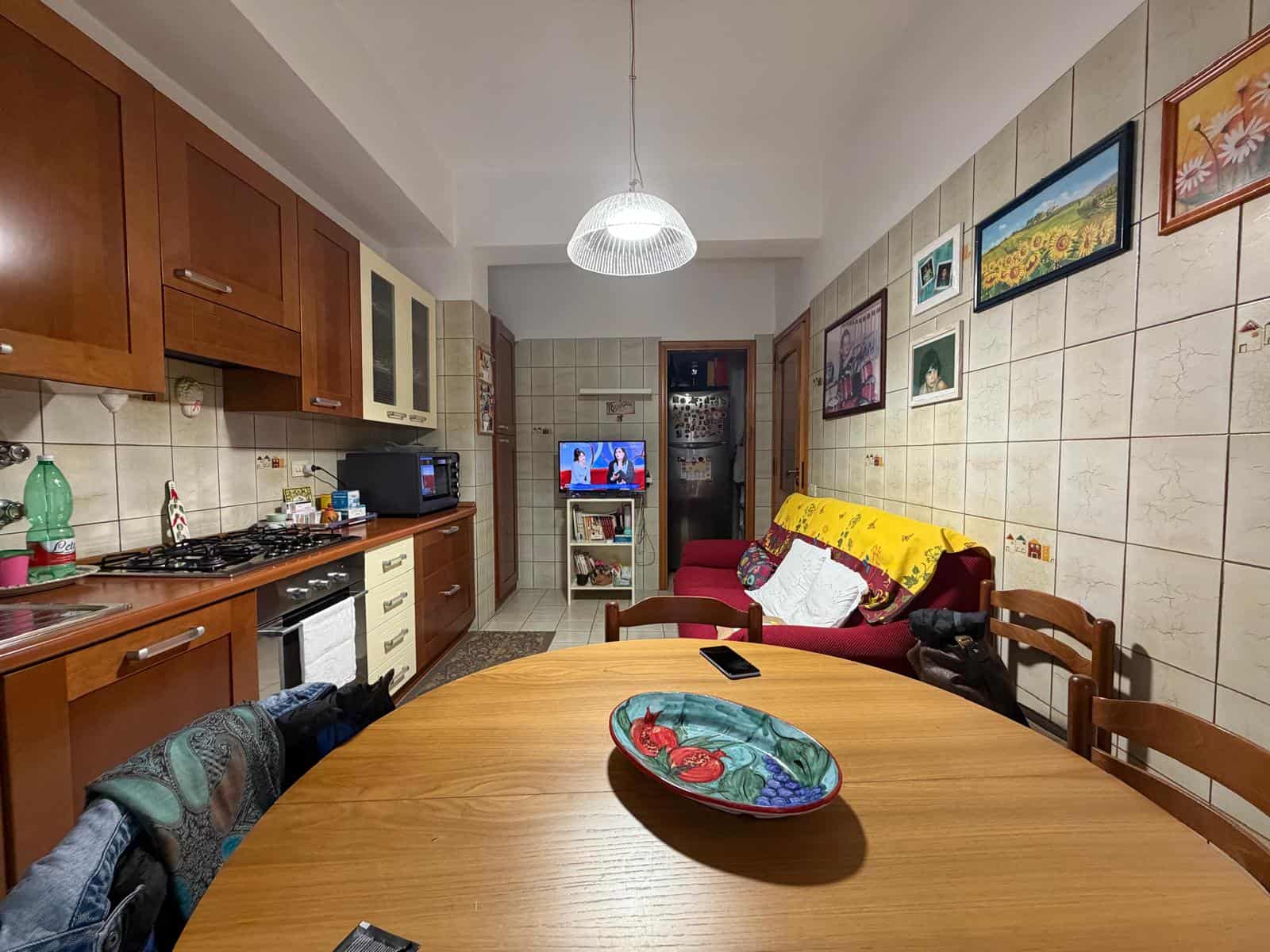 Lavagna di cucina con soggiorno, mobili in legno, pareti decorate con quadri colorati, tavolo rotondo con piatto decorativo, ambiente accogliente e luminoso.