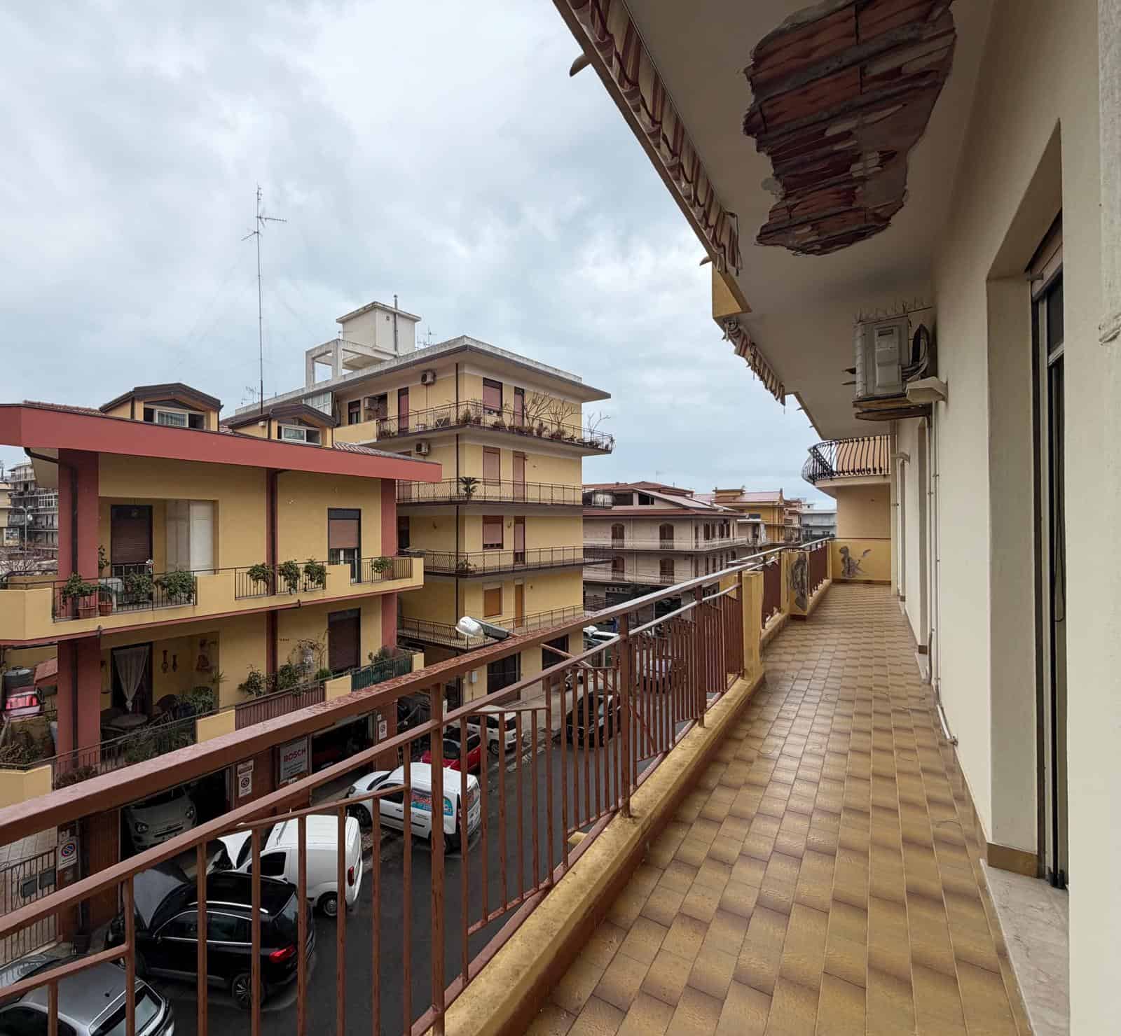 Balcone di un appartamento in un condominio urbano con vista sulle altre abitazioni e sulle auto parcheggiate sotto, cielo nuvoloso.