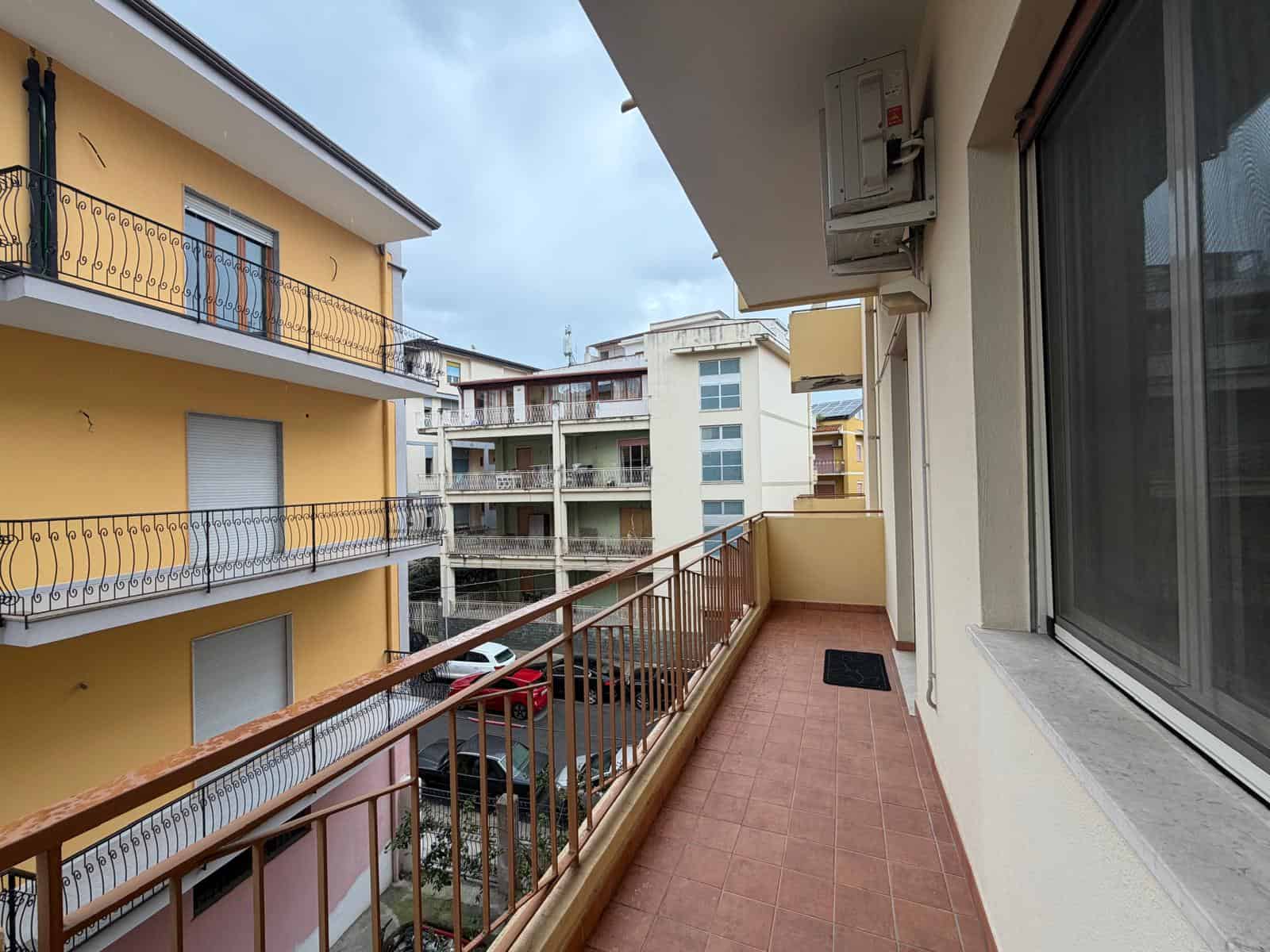 Balcone di un appartamento in un complesso residenziale, vista su edifici moderni e parcheggio sottostante, con pavimentazione in piastrelle e ringhiera in metallo, in un contesto urbano.