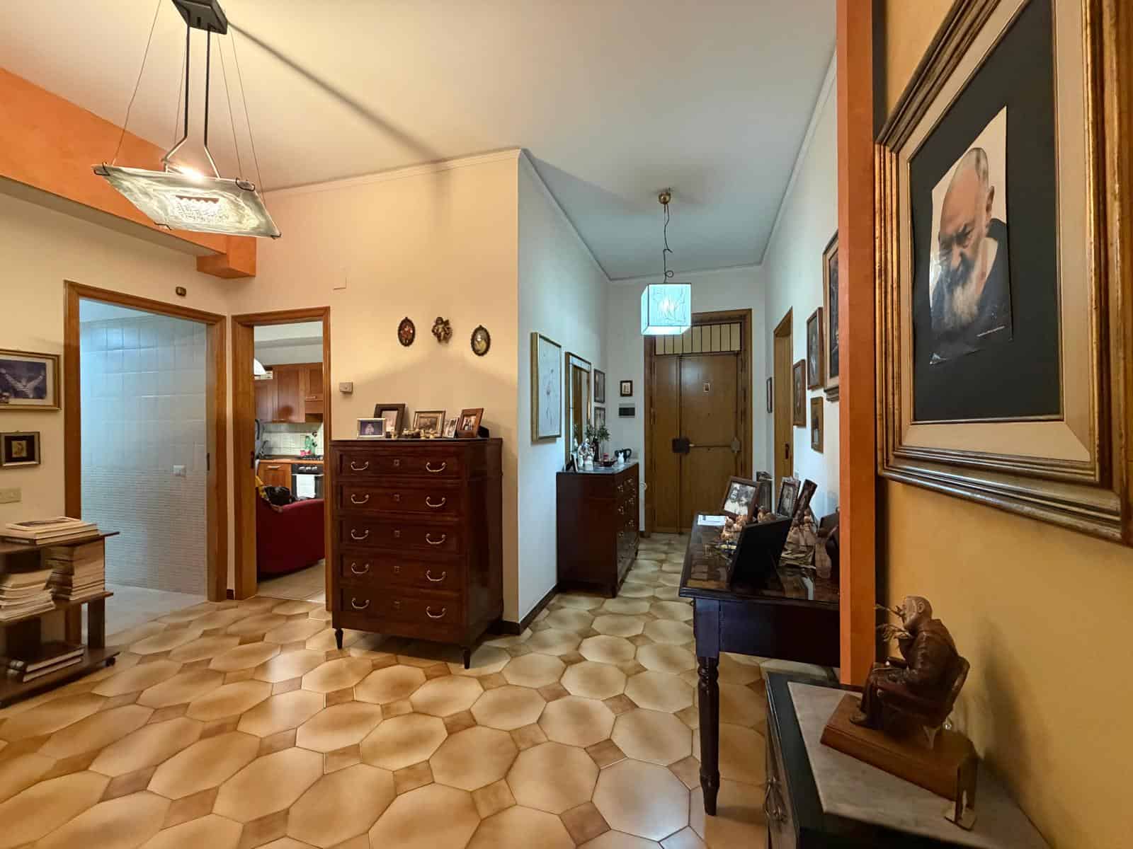 Una vista dell'ingresso di una casa con pareti decorate, mobili in legno e quadri alle pareti, illuminata da luci soffuse e lampade da soffitto, creando un ambiente accogliente e elegante.