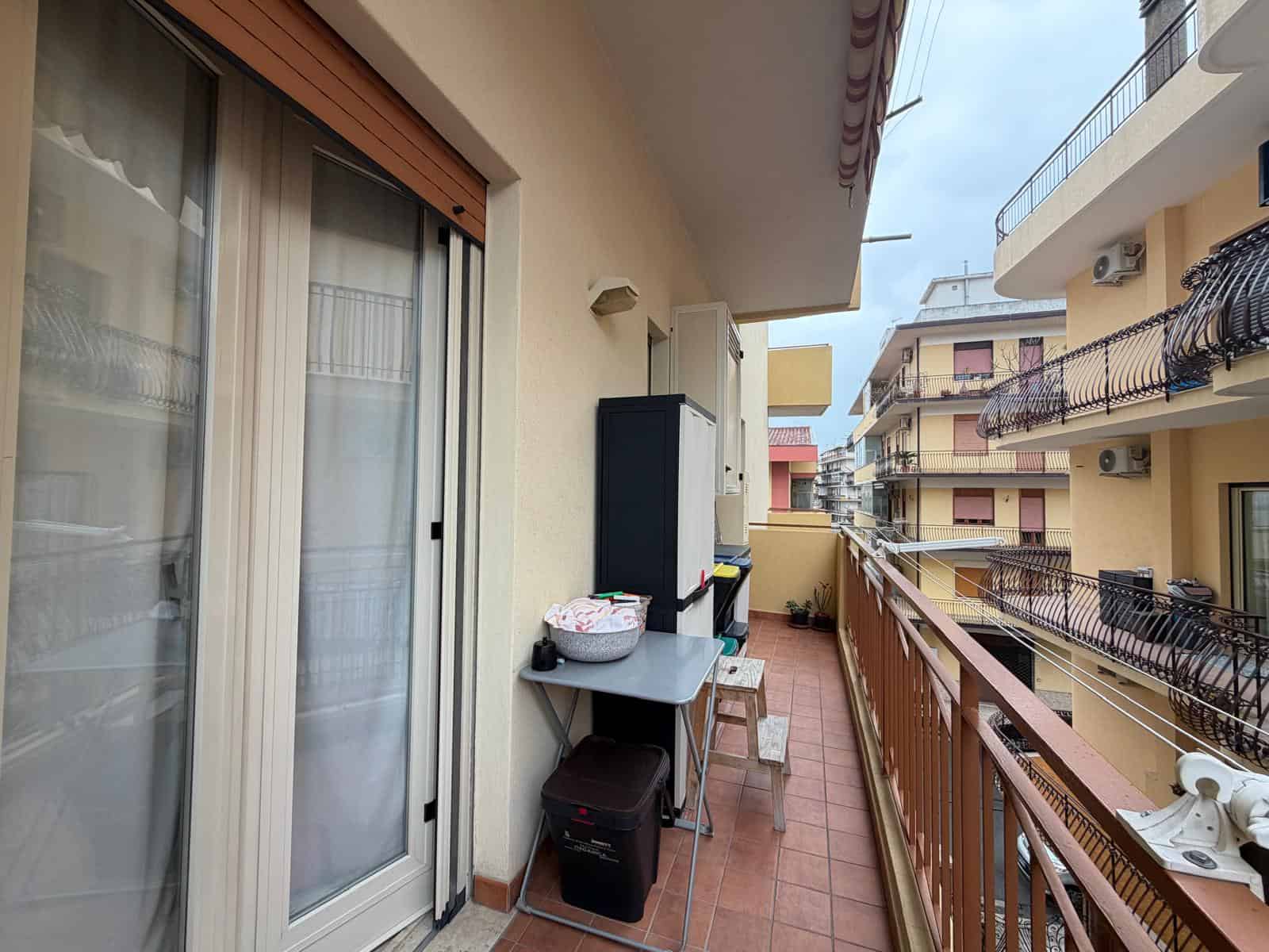Ampio balcone di un appartamento con vista su edifici residenziali, con mobili da esterno e piante, ideale per relax e socializzazione all'aperto.