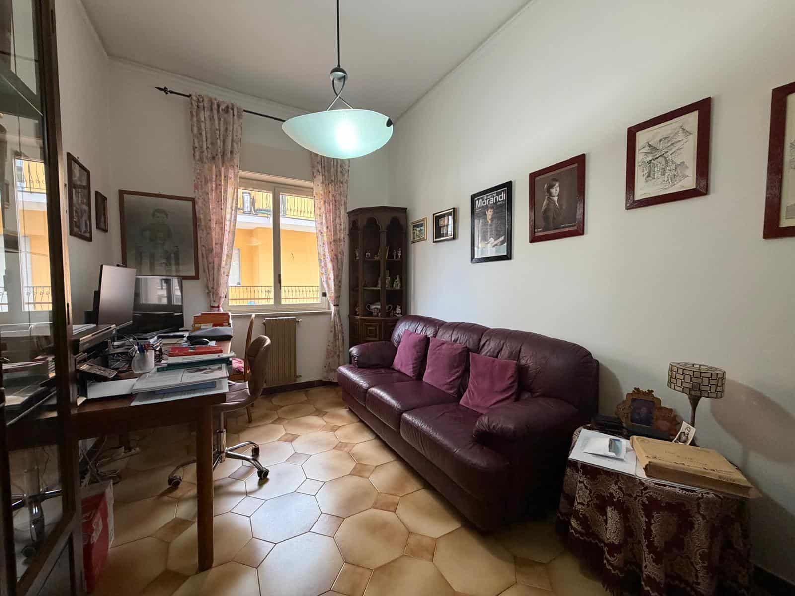 Interno di un soggiorno con divano in pelle, scrivania con computer e pareti decorate con quadri, ambiente luminoso e accogliente.