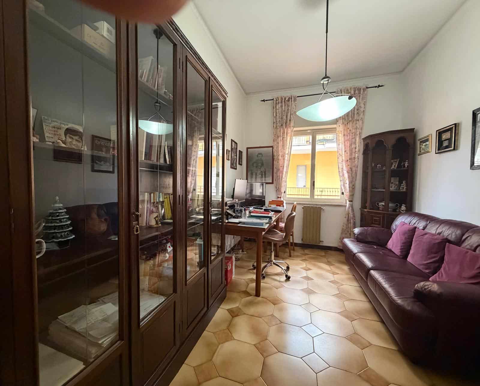 Arredo ufficio domestico con scrivania, sedia e libreria in stile classico, illuminato da luce naturale e lampada moderna, in ambiente accogliente e ben organizzato.