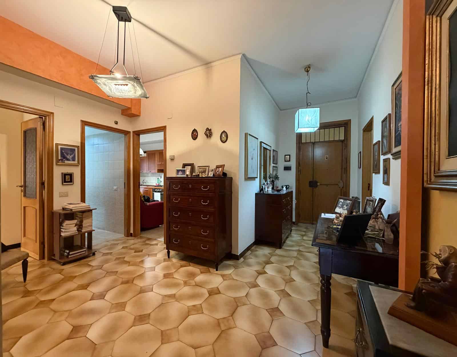 Una vista dell'ingresso di una casa con pavimento in piastrelle beige, pareti bianche e arancioni, e mobili in legno, con quadri e decorazioni alle pareti, illuminata da lampade moderne e tradizionali.