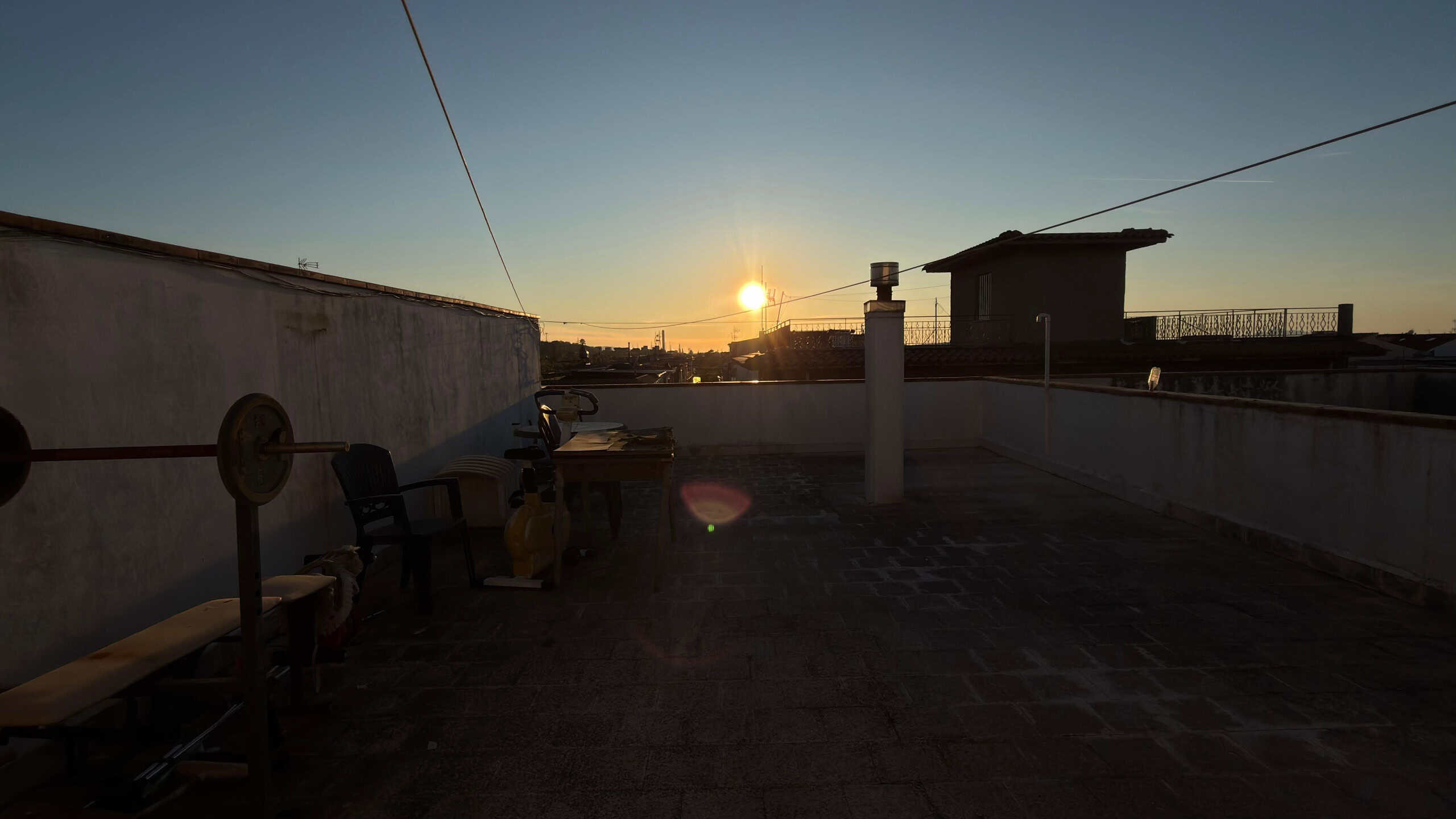 Terrazza sul tetto con vista al tramonto, sedie e attrezzature per il fitness, in un ambiente urbano, con il sole che tramonta all'orizzonte.