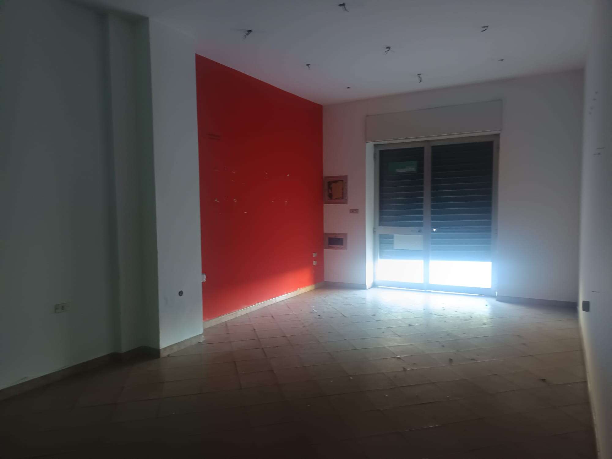 Aggiunta di un'immagine di un interno di appartamento con parete rossa e grande finestra con tapparelle, pronto per essere arredato e personalizzato.