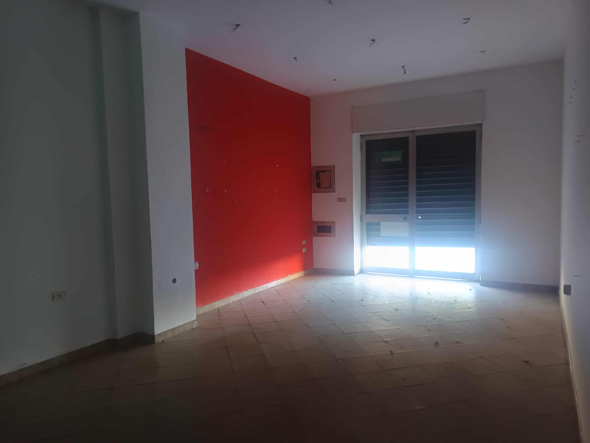 Aggiunta di un'immagine di un interno di appartamento vuoto con pareti bianche e una parete rossa, grande finestra con tapparelle e pavimento in parquet.