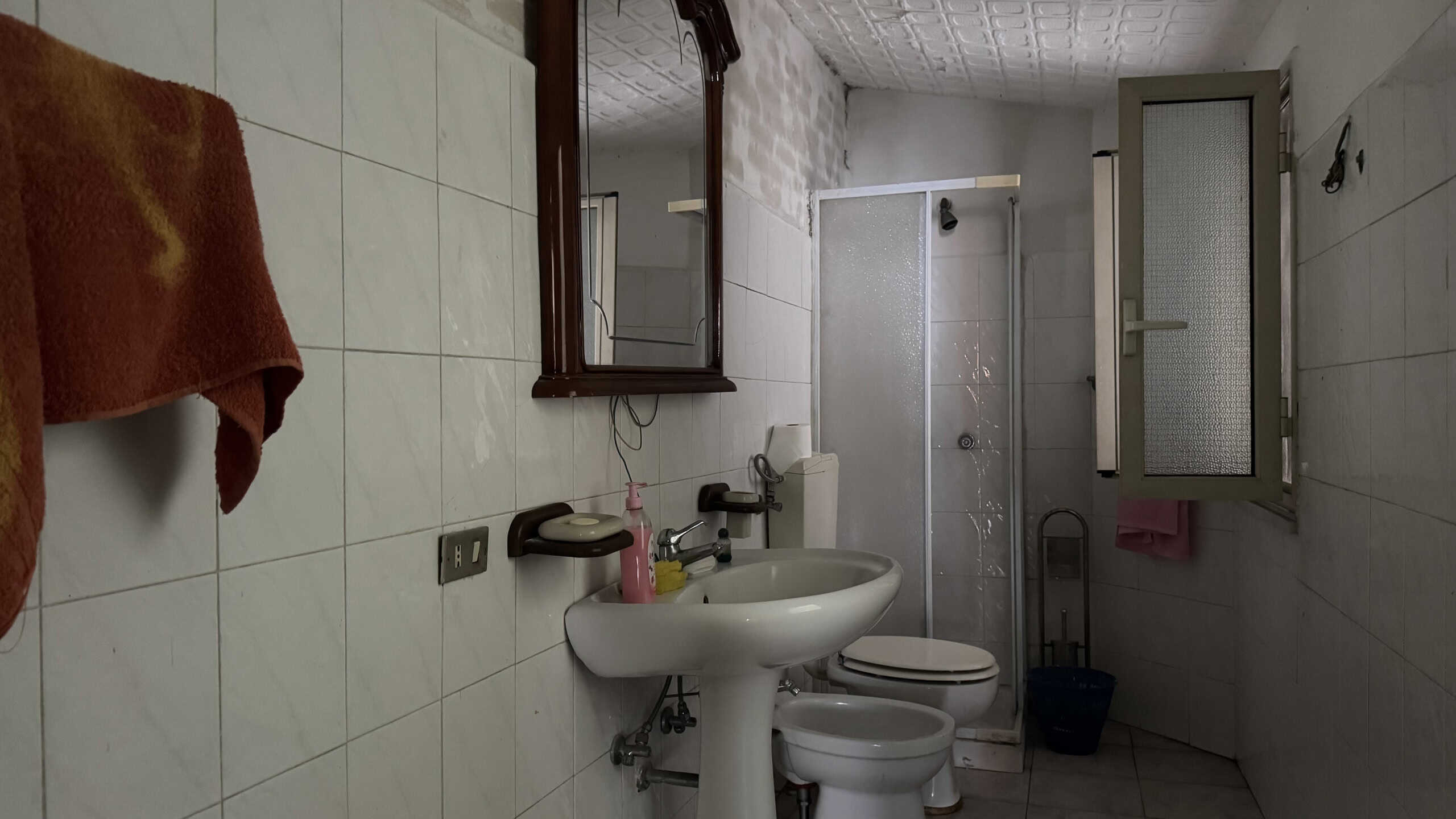 Lavabo e servizi igienici in bagno con piastrelle bianche, specchio e finestra per ventilazione naturale, ambiente funzionale e ben illuminato.