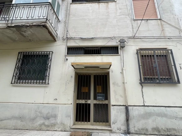 Ingresso di un edificio residenziale con porta in metallo e finestre con grate di sicurezza, pareti in cemento e balcone con ringhiera in ferro, in zona urbana.
