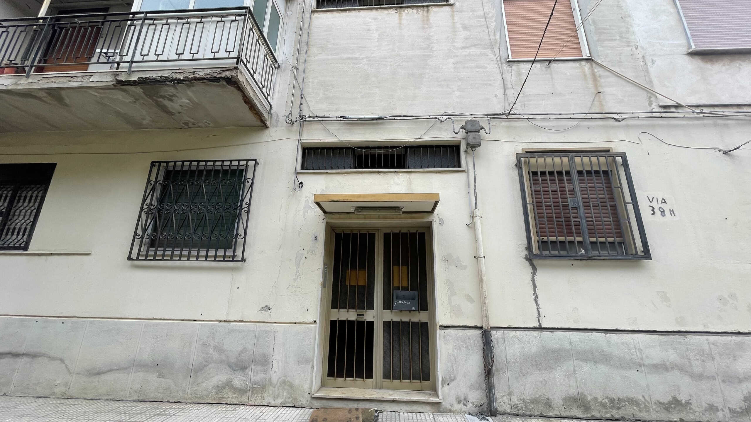 Ingresso di un edificio residenziale con porta in metallo e finestre con grate di sicurezza, pareti in cemento e balcone con ringhiera in ferro, in zona urbana.