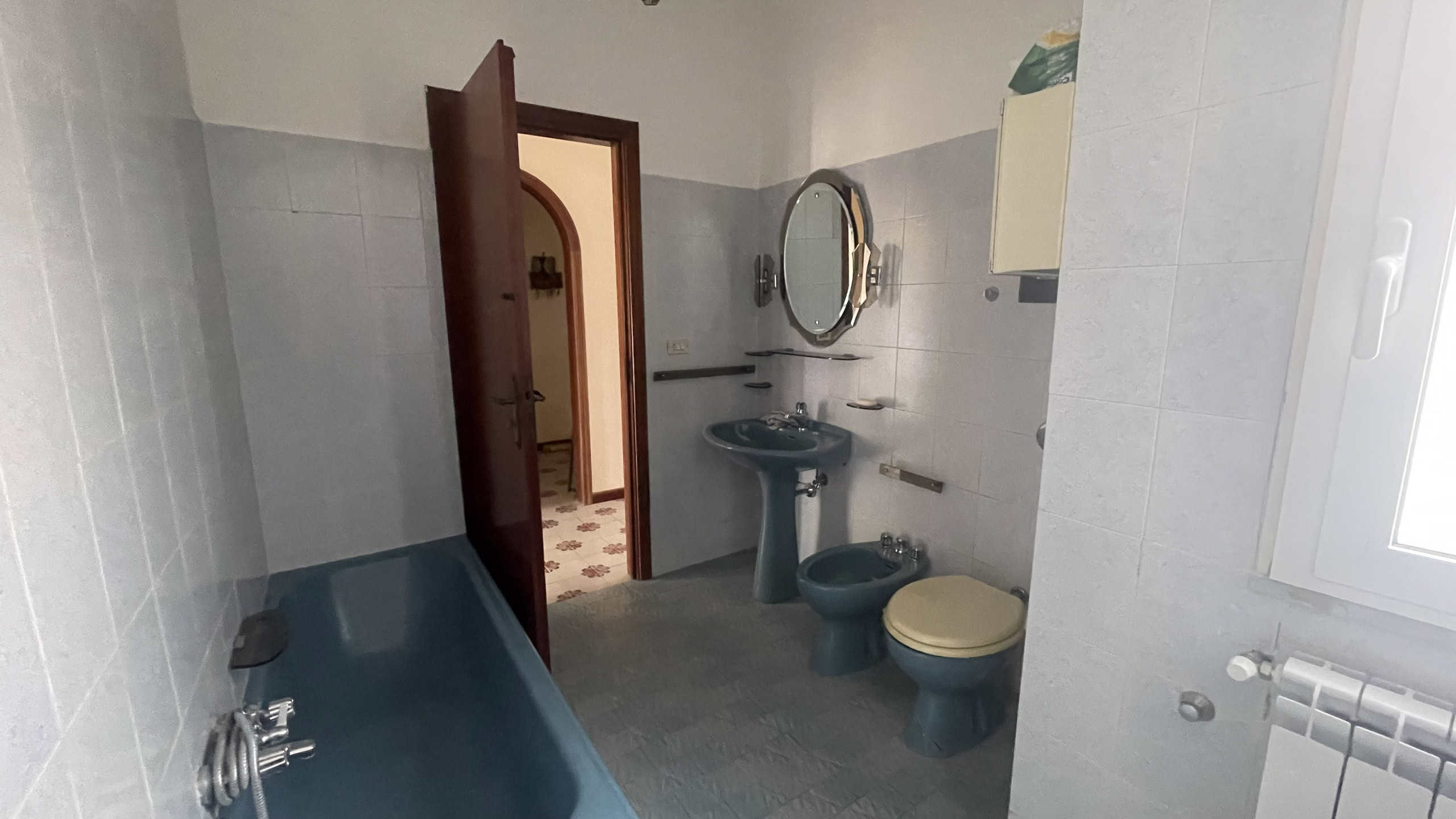 Lavabo e servizi igienici in bagno con piastrelle grigie, specchio rotondo e finestra per ventilazione naturale, ambiente funzionale e ben illuminato.
