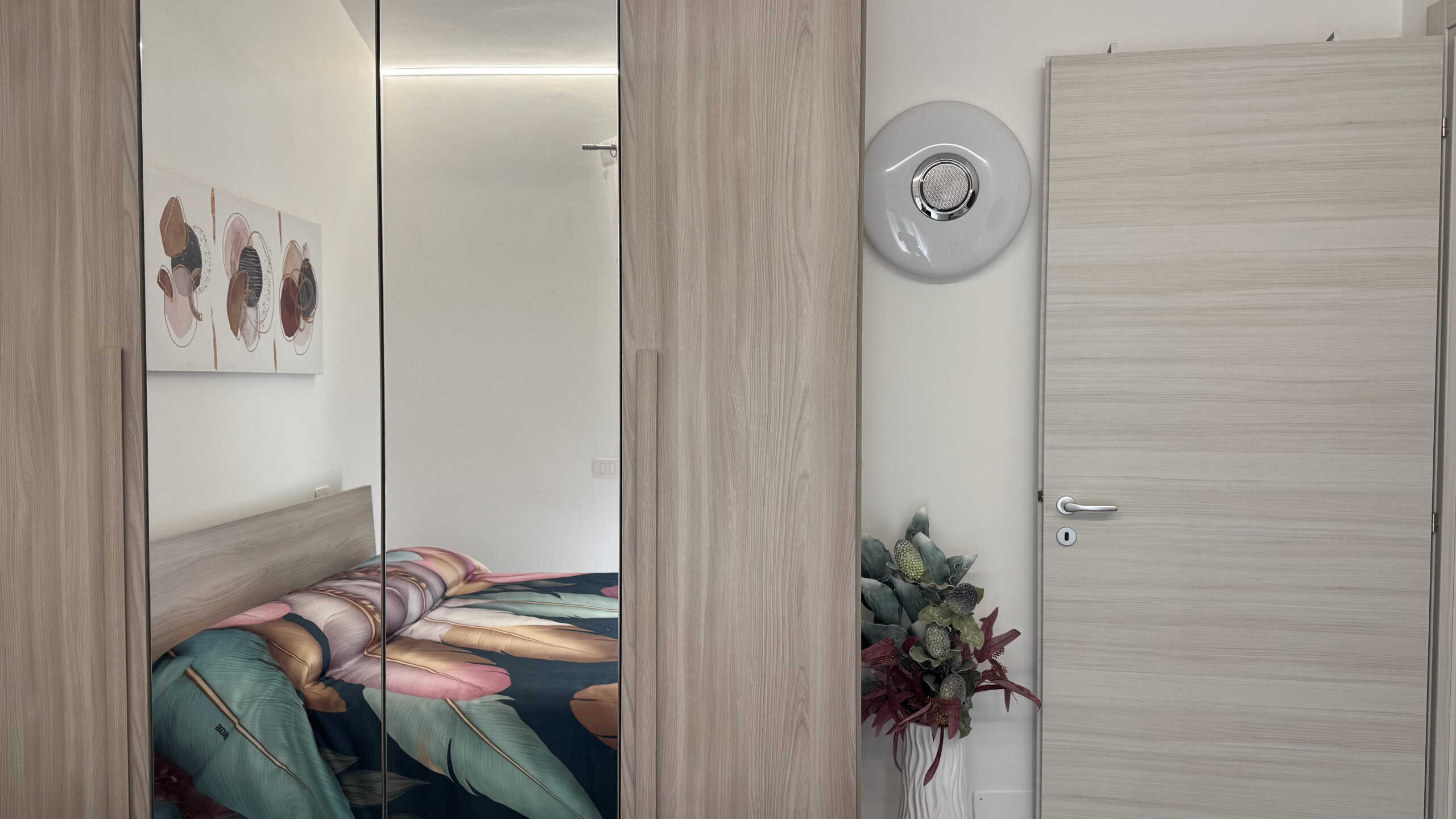 Elegante camera da letto moderna con arredi in legno chiaro, letto con biancheria colorata e decorazioni artistiche alle pareti, ambiente luminoso e accogliente.