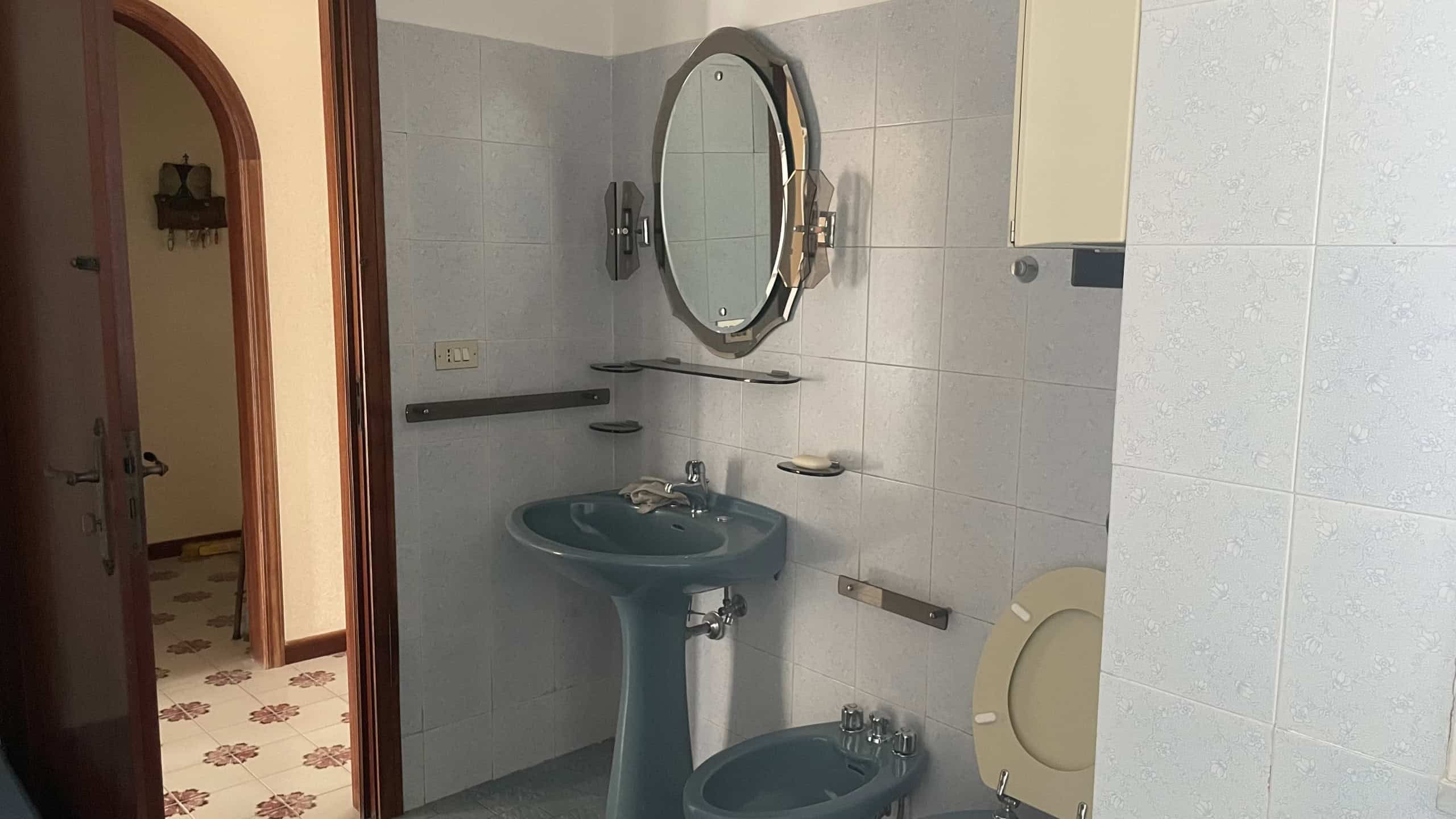 Specchio rotondo e lavabo in bagno con pareti rivestite di piastrelle grigie, accessori e maniglie in metallo, ambiente pulito e funzionale.