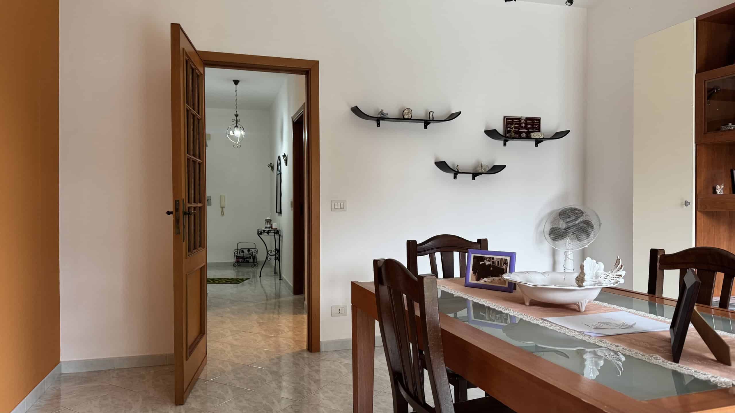 Elegante soggiorno con tavolo in legno e sedie, pareti bianche e decorazioni moderne, ideale per vivere momenti di relax e convivialità in un ambiente accogliente.