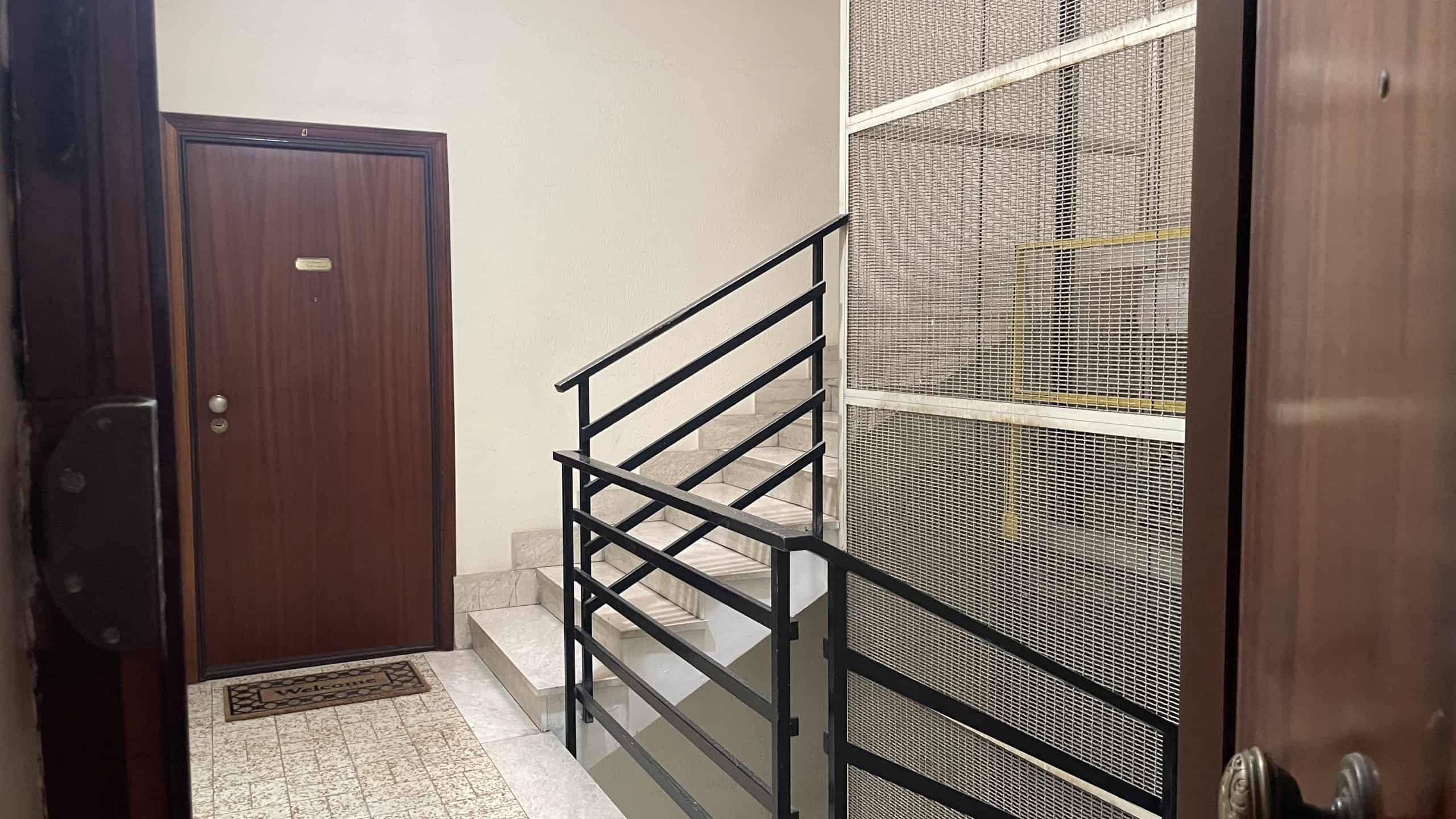 Escalator in un edificio residenziale con ringhiera in metallo nero e pareti in legno, ingresso con porta in legno e tappetino.