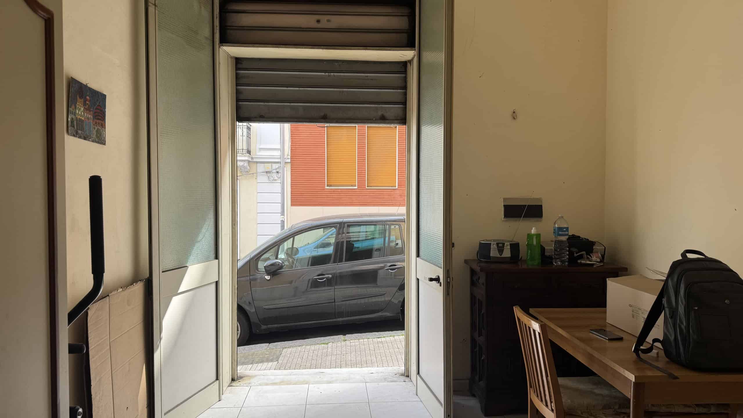 Ingresso di un appartamento con vista sulla strada e un'auto parcheggiata, ingresso con porta aperta, ambiente interno con scrivania e oggetti vari, illuminato dalla luce naturale.