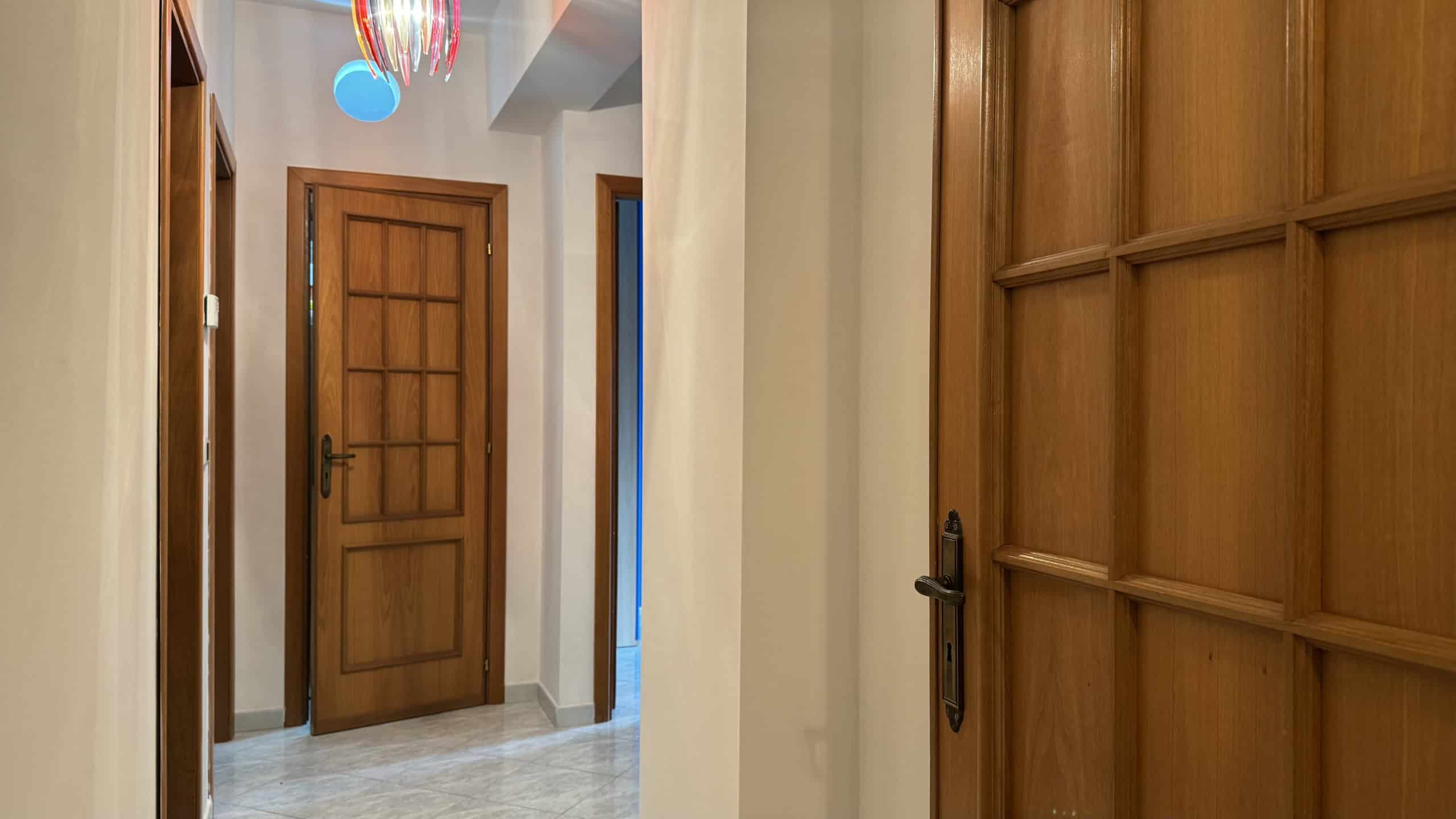 Ampia porta in legno massiccio in un ingresso di casa, con dettagli eleganti e maniglia nera, illuminata da luce naturale e decorazioni colorate sul soffitto.