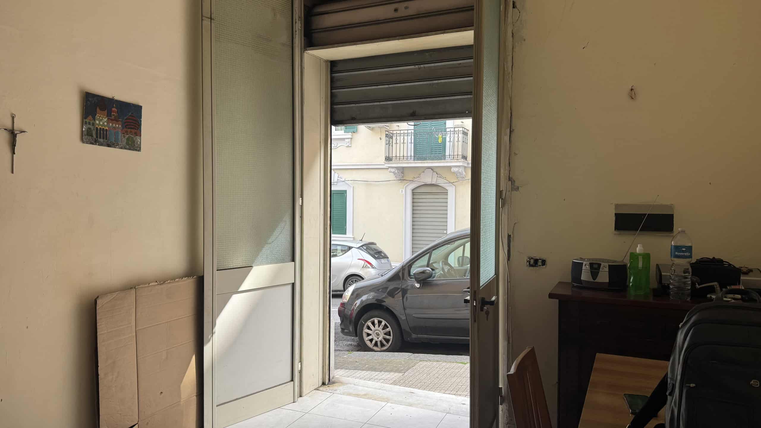 Ampio ingresso con porta vetrata che dà su strada, con vista su auto parcheggiate e edifici storici, illuminato dalla luce naturale, ideale per un ambiente residenziale o commerciale.