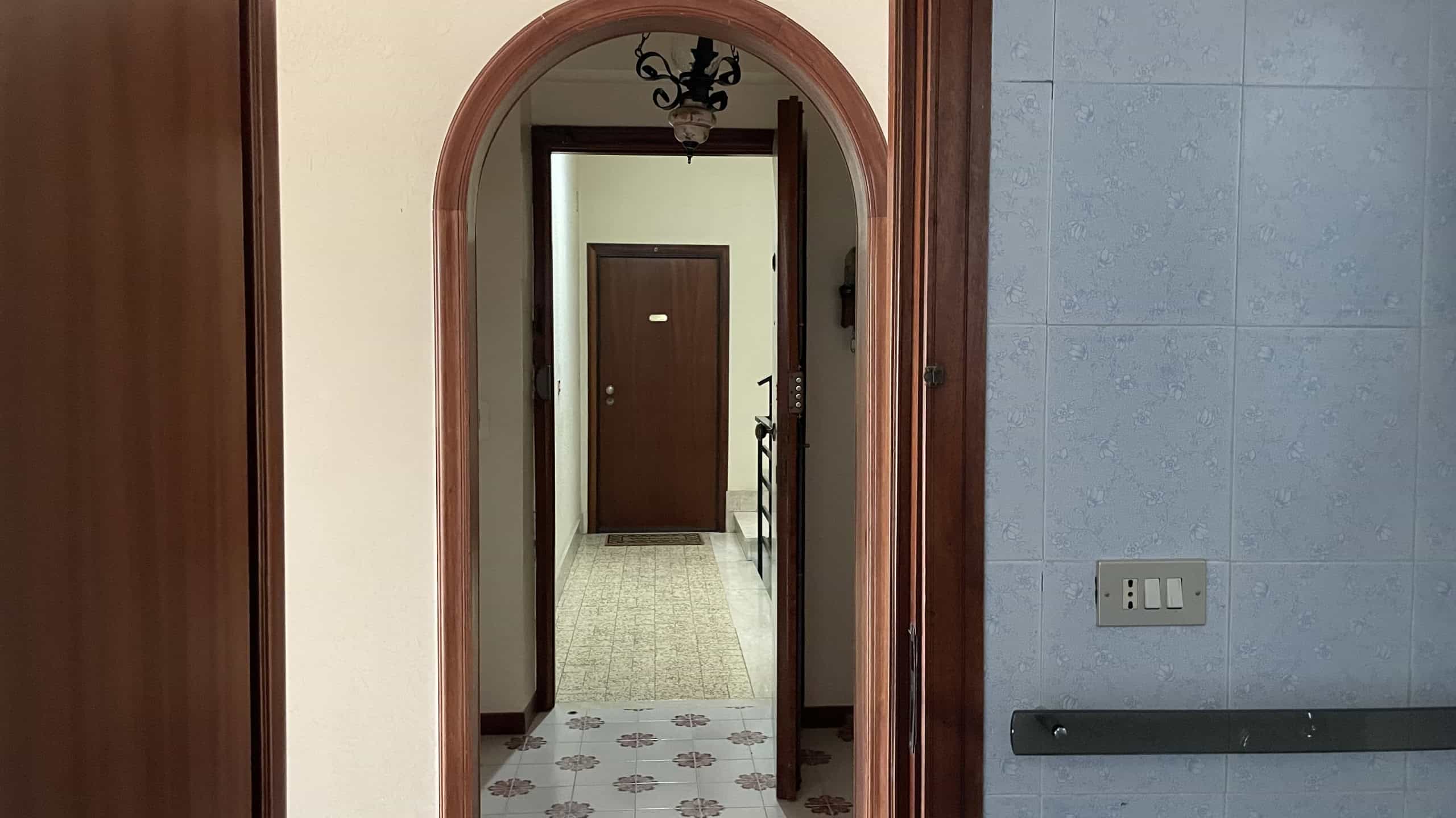 Ampia porta d'ingresso in legno con arco, vista da un corridoio interno con pavimento in piastrelle e pareti chiare, illuminato da luce naturale e lampadario decorativo.