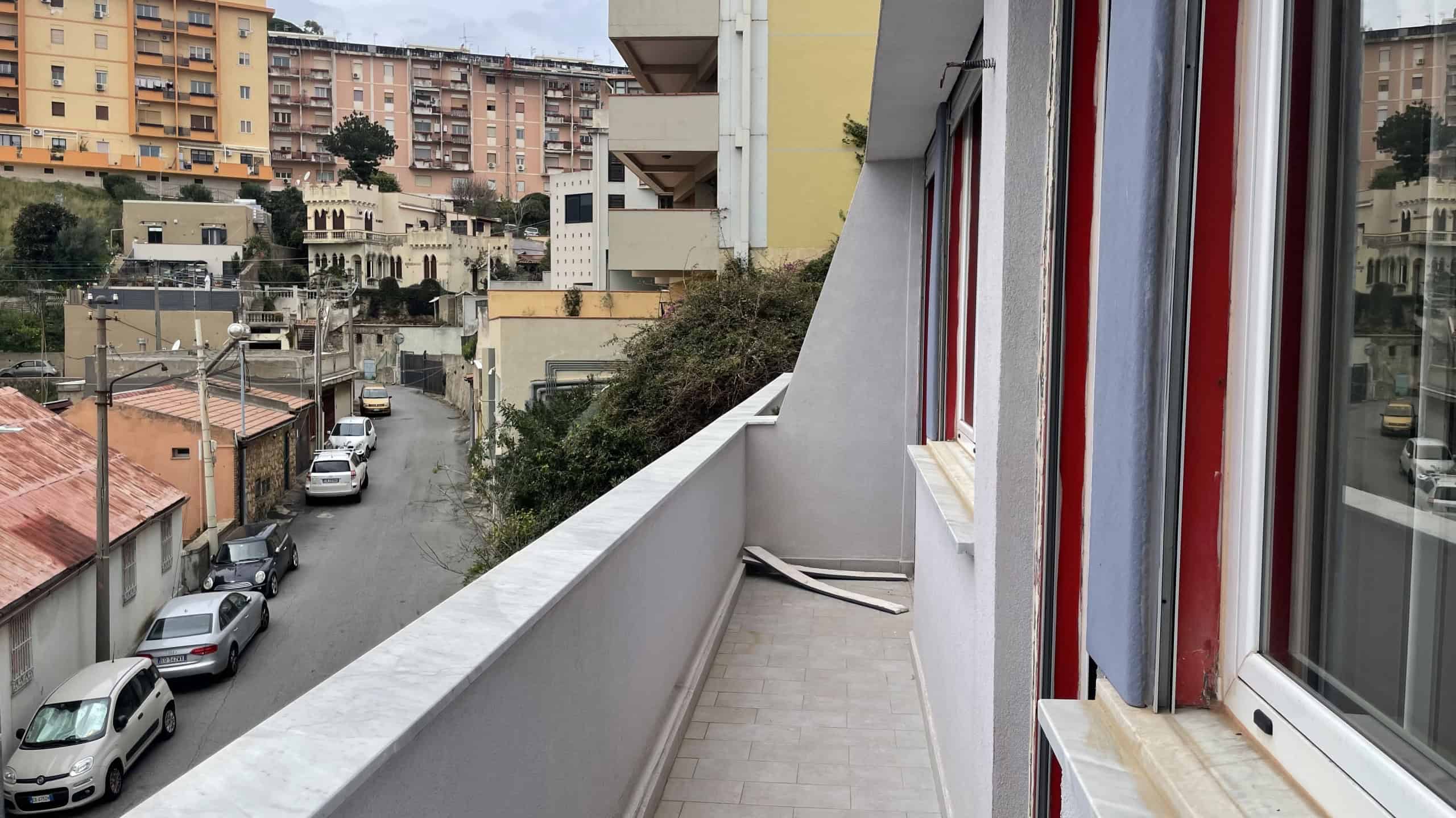 Balcone di un appartamento con vista urbana, pavimentato e con pareti bianche, situato in un quartiere residenziale di città, con edifici alti e strade trafficate visibili in lontananza.
