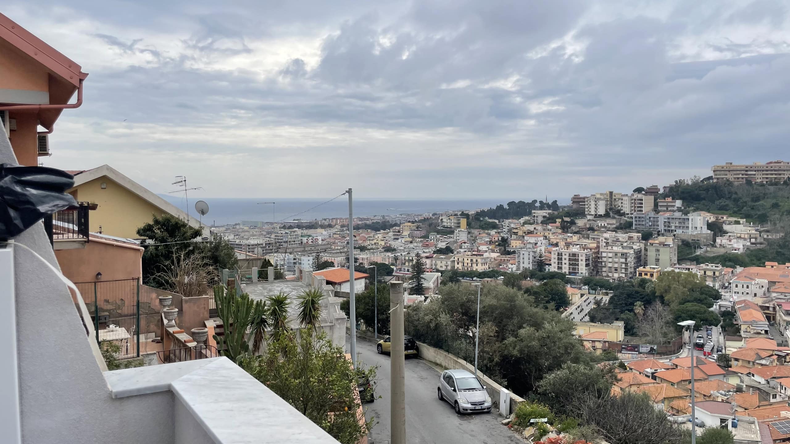 Vista panoramica sulla città di Napoli con il mare sullo sfondo, case colorate e colline verdi, sotto un cielo nuvoloso, rappresentando un paesaggio urbano mediterraneo.