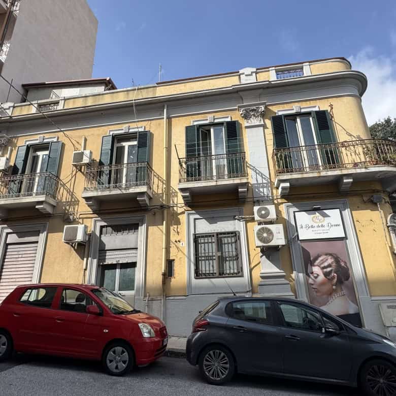 Edificio storico in stile classico con balconi in ferro battuto e facciata gialla, situato nel centro di Napoli, con auto parcheggiate davanti e cielo sereno.