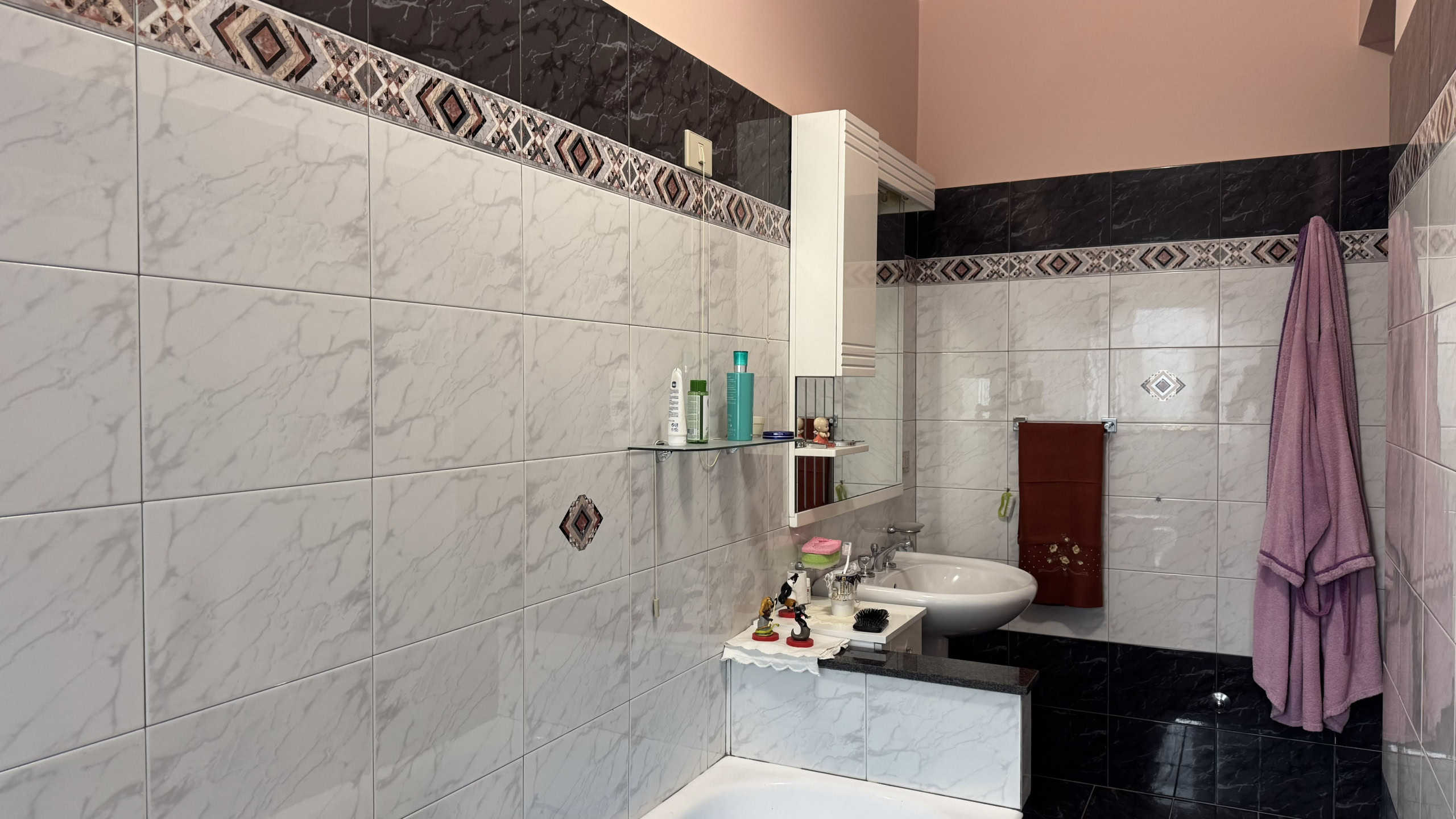 Lavabo moderno in bagno con piastrelle bianche e dettagli decorativi, accessori e asciugamano viola appeso alla parete.