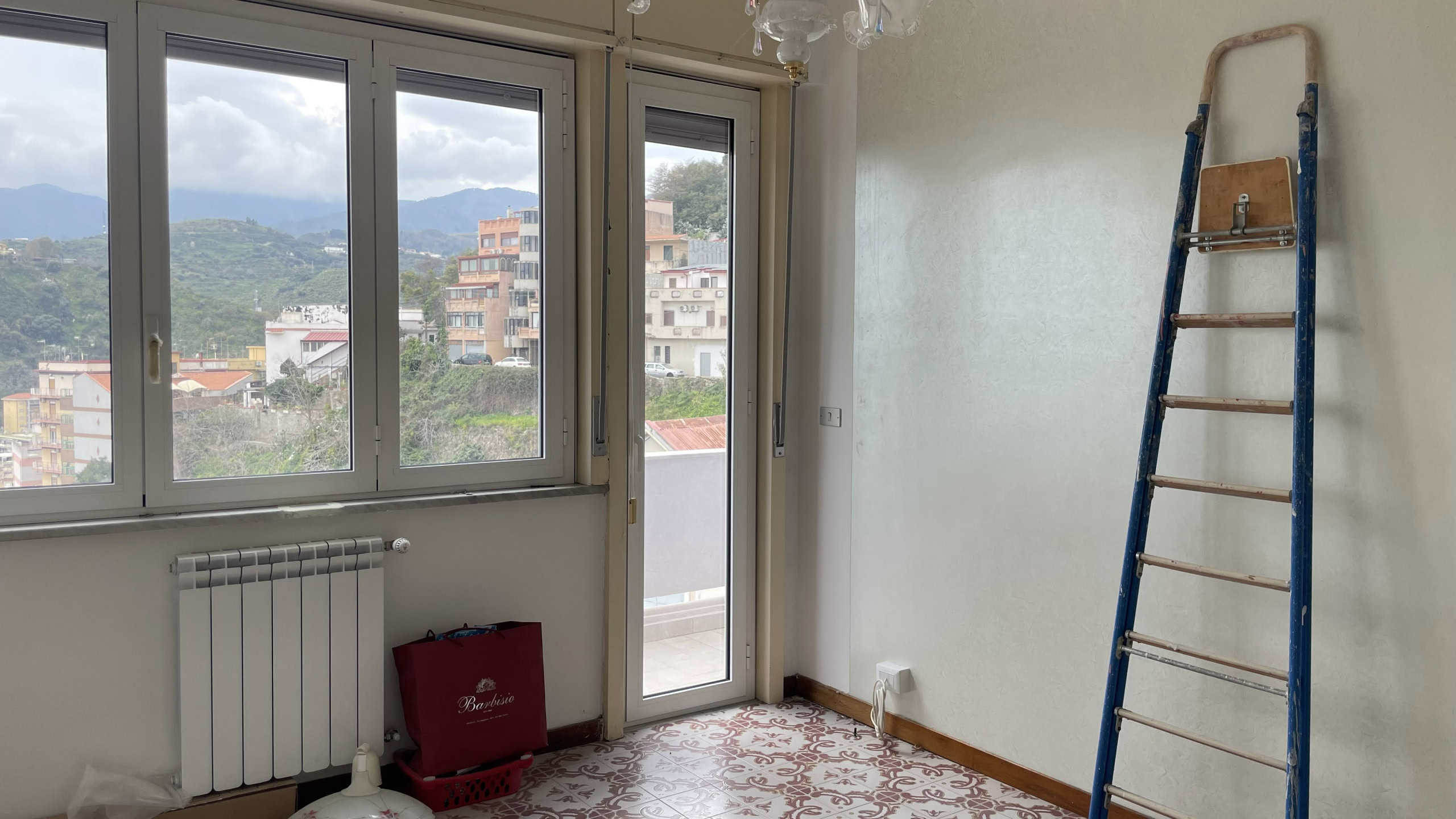 Serramenti in alluminio con vista panoramica sulla città e sulle montagne, luce naturale che illumina l'interno di una stanza in fase di ristrutturazione con scala a pioli e pavimento in parquet.