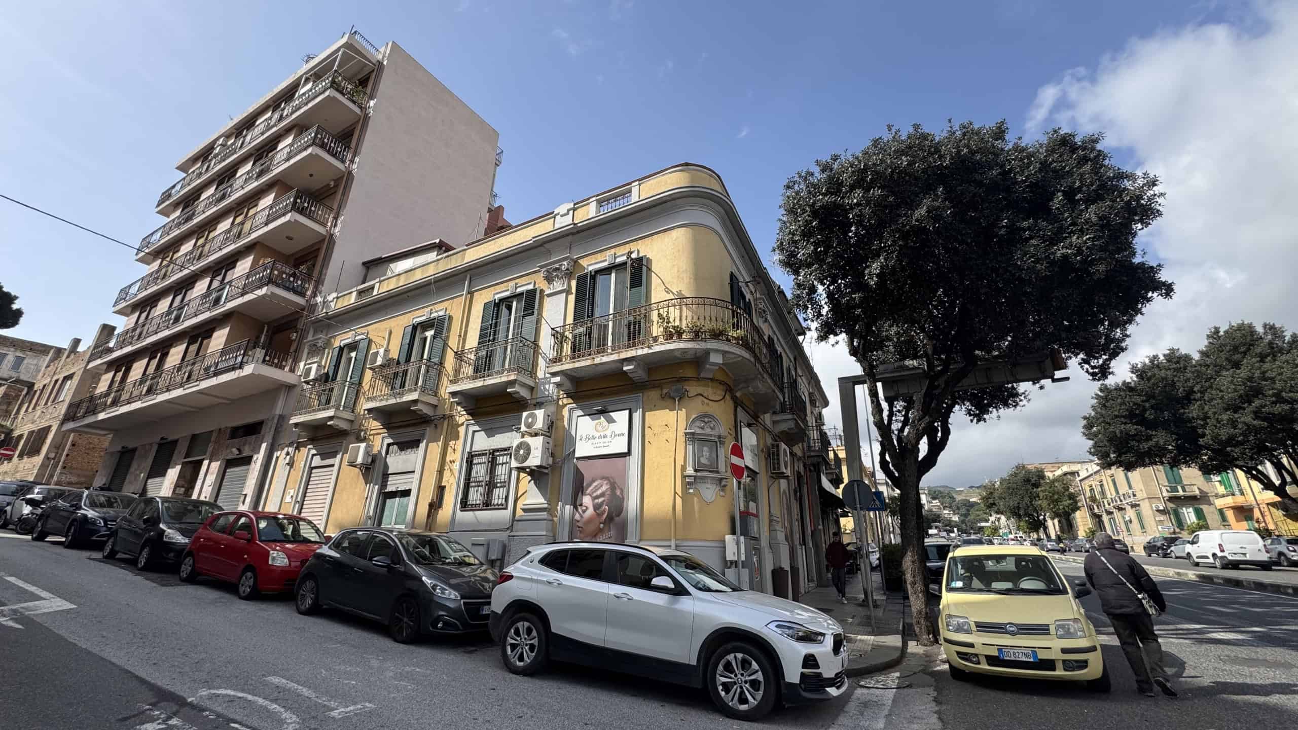 Elegante edificio storico con facciata gialla e balconi in ferro battuto, situato in una zona urbana con auto parcheggiate e alberi, rappresenta un esempio di architettura residenziale in centro città.