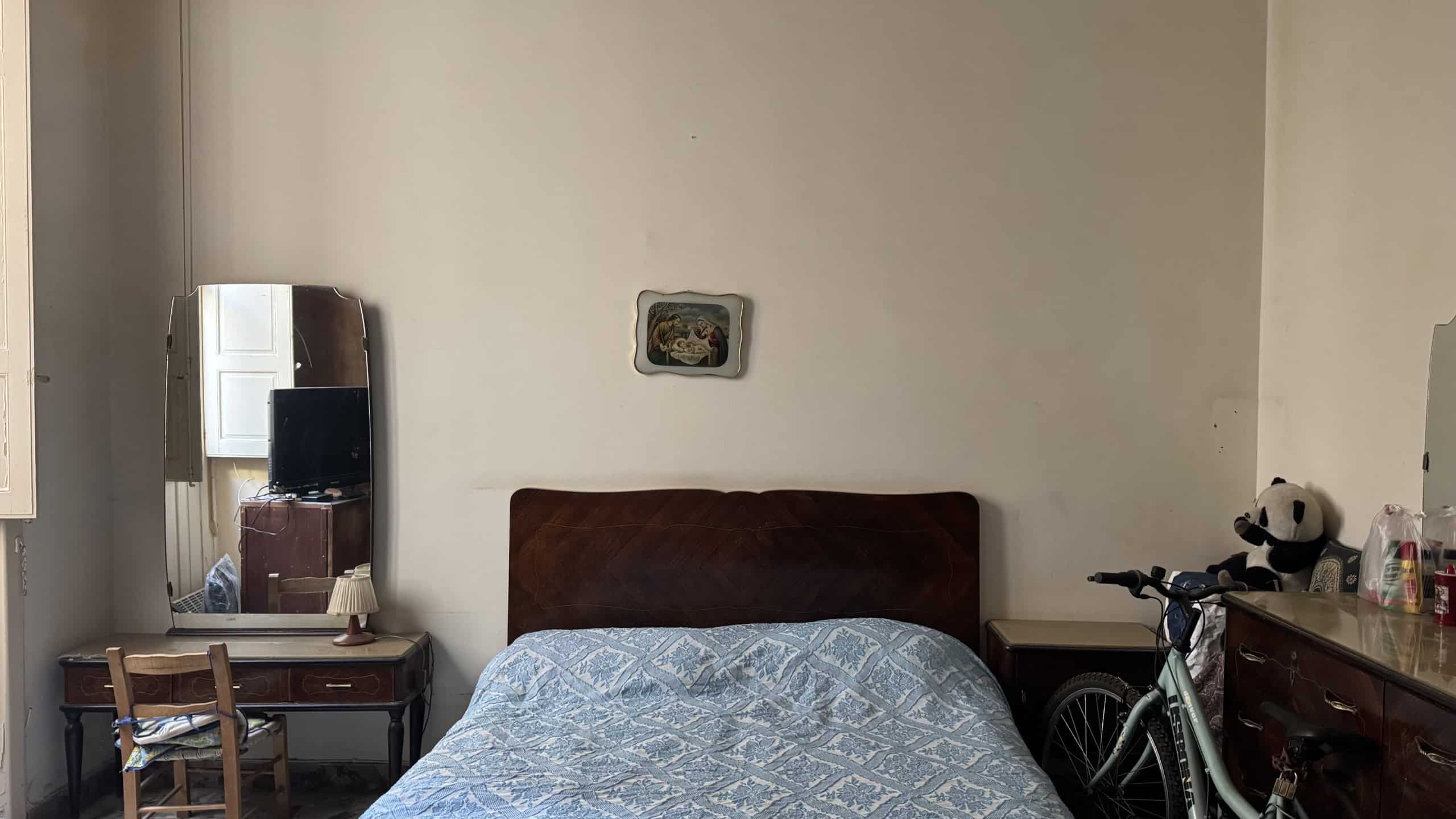 Letto matrimoniale con testiera in legno scuro, in una stanza luminosa con pareti chiare, decorata con un quadro sopra la testata. Presente un mobile con oggetti e una bicicletta vicino al letto.