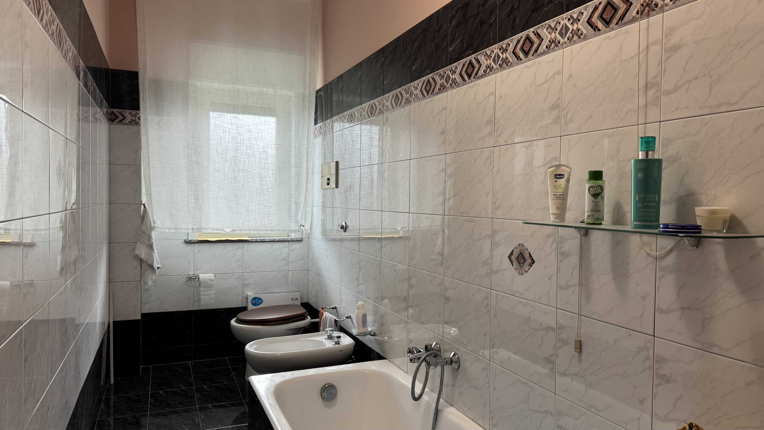 Bagno moderno con vasca, bidet e lavabo, pareti rivestite in piastrelle bianche e dettagli decorativi, finestra con tende leggere, ideale per appartamenti in vendita o affitto a NextCasa.