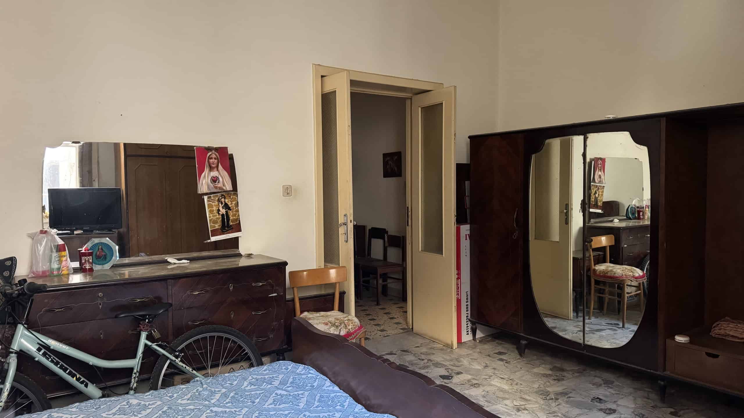 Armadio a specchio e mobili in stile vintage in una camera da letto con pareti beige e pavimento in piastrelle. Spazio organizzato con mobili funzionali e decorazioni semplici, ideale per ambienti res.