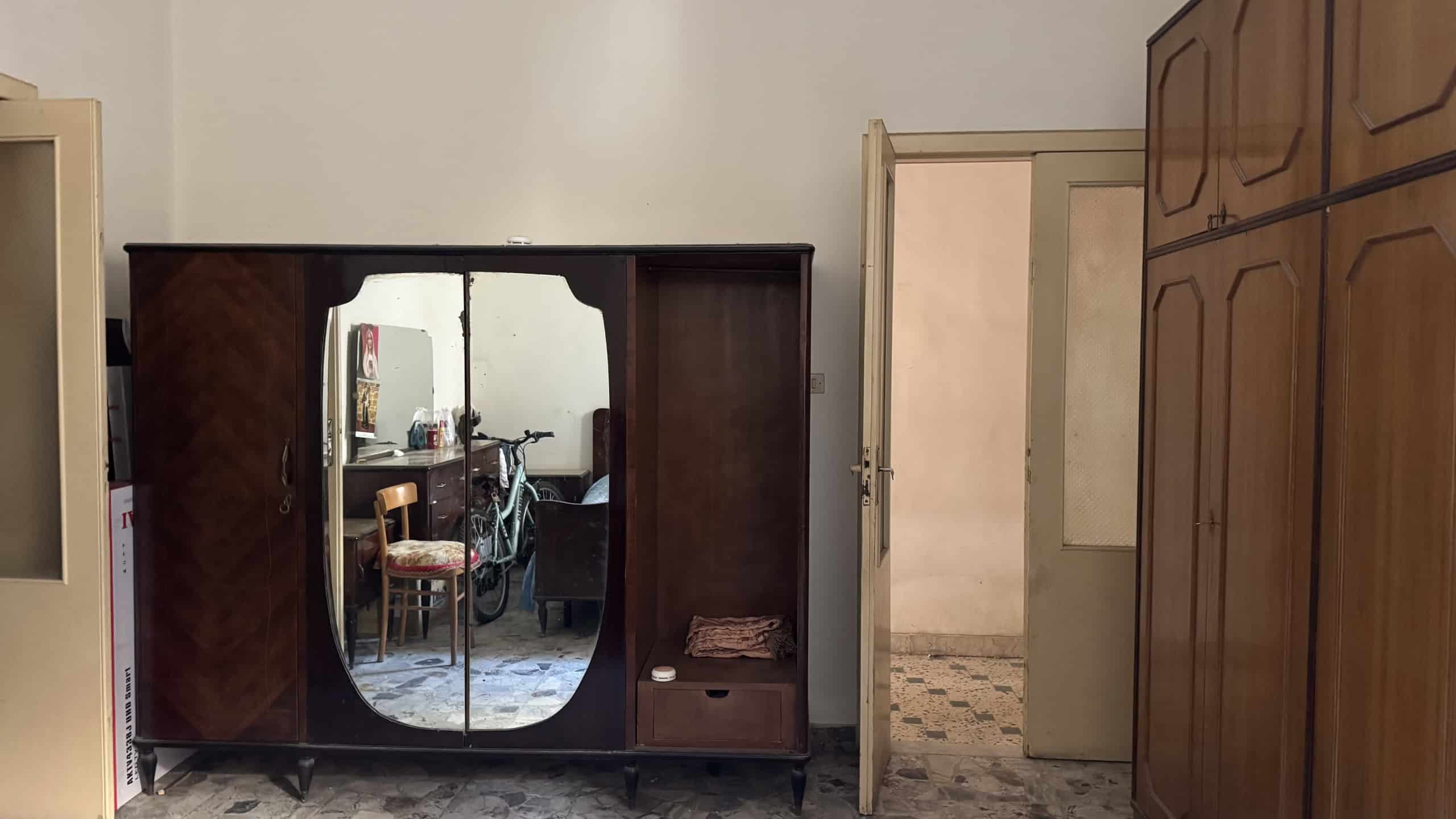 Armadio in legno scuro con specchi, in un ambiente domestico, con porta aperta che rivela una stanza adiacente. Ideale per arredamento classico e funzionale, perfetto per ottimizzare lo spazio in casa.
