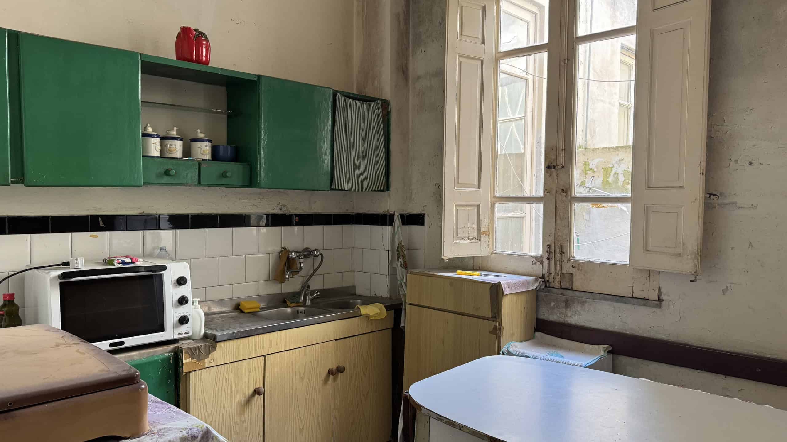 Lavandino e cucina in stile vintage con ante verdi e pareti in piastrelle bianche, illuminata dalla luce naturale delle finestre con persiane in legno.