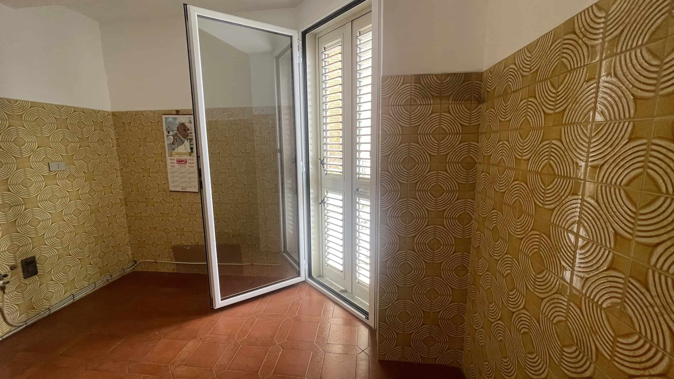 Ampia stanza con pareti rivestite in piastrelle gialle con motivi geometrici, porta finestra che permette l'accesso a un balcone o terrazzo, illuminata dalla luce naturale.