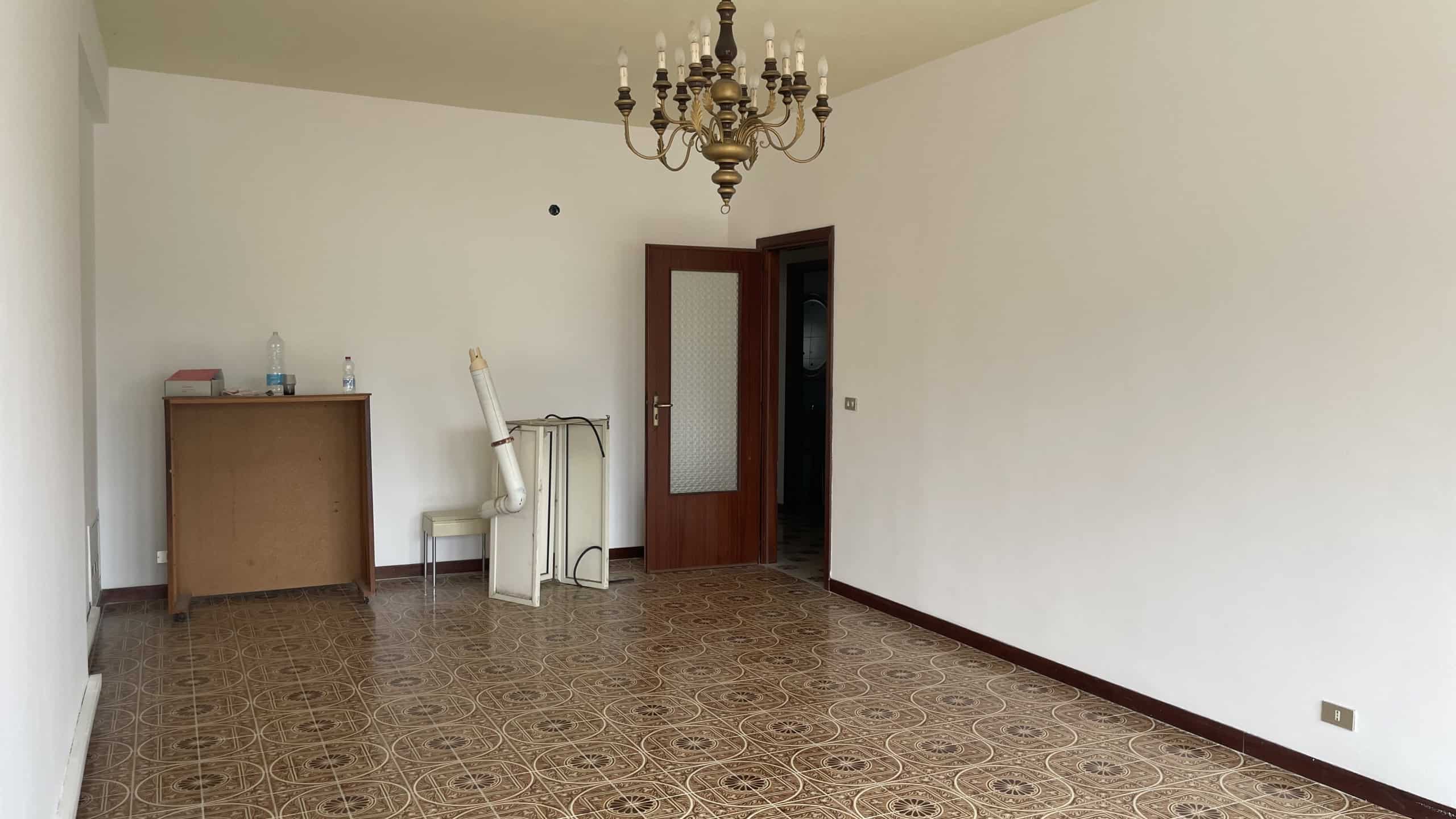 Appartamento vuoto con pareti bianche, pavimento in piastrelle decorative e lampadario antico, pronto per essere arredato e personalizzato.