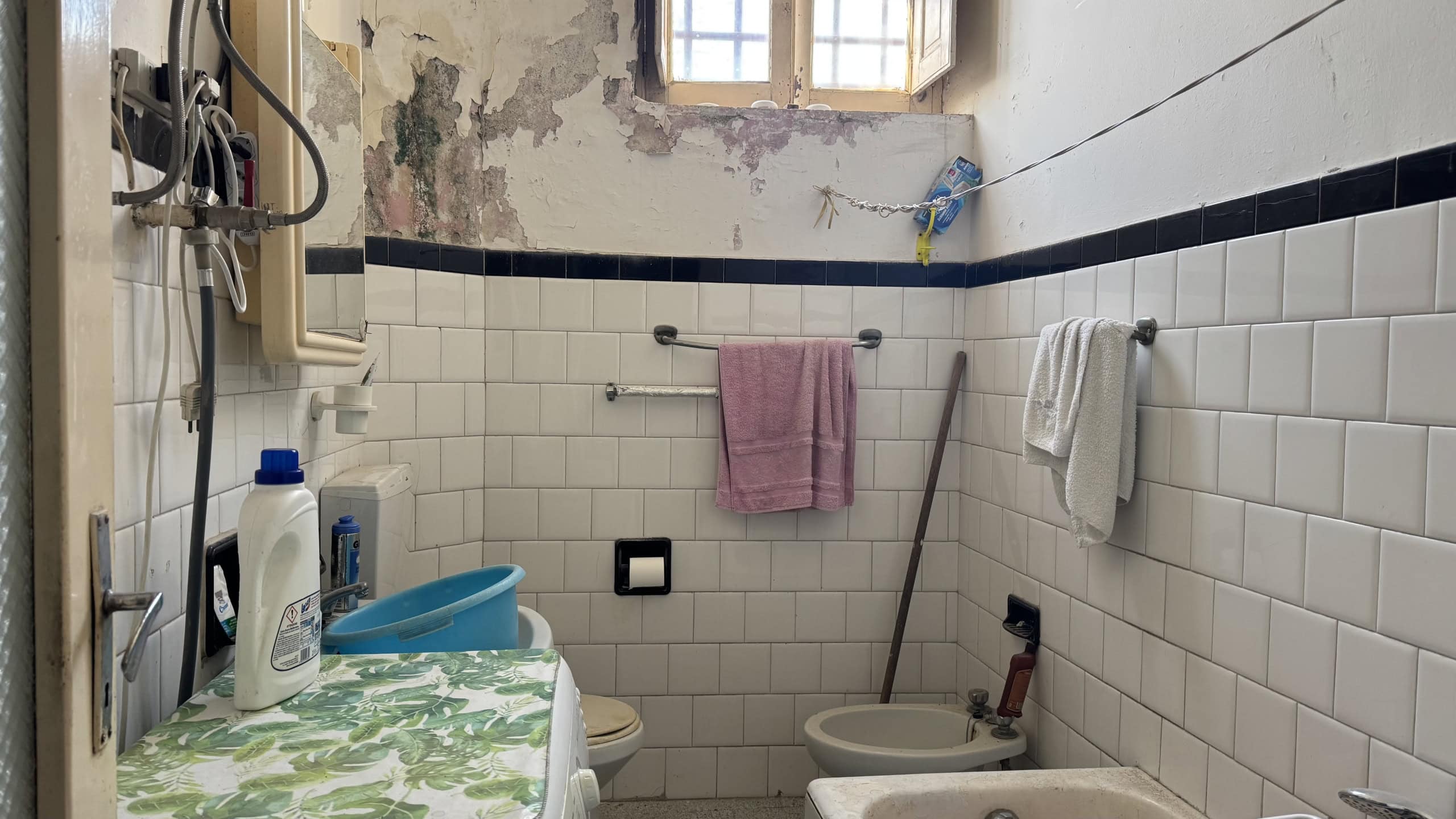 Spazzatura e pareti danneggiate in un bagno abbandonato, con piastrelle bianche e accessori di base, evidenziando condizioni di degrado e abbandono.