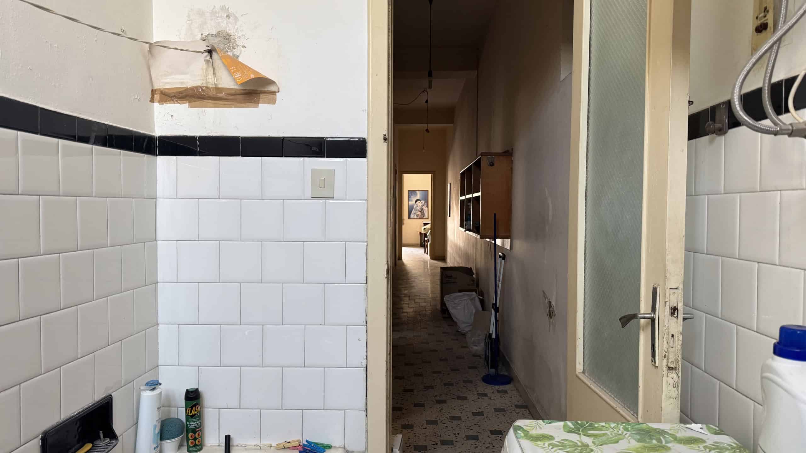 Ampia vista di un corridoio interno di un appartamento con pareti rivestite di piastrelle bianche e dettagli neri, con porta aperta che conduce a un'altra stanza luminosa.