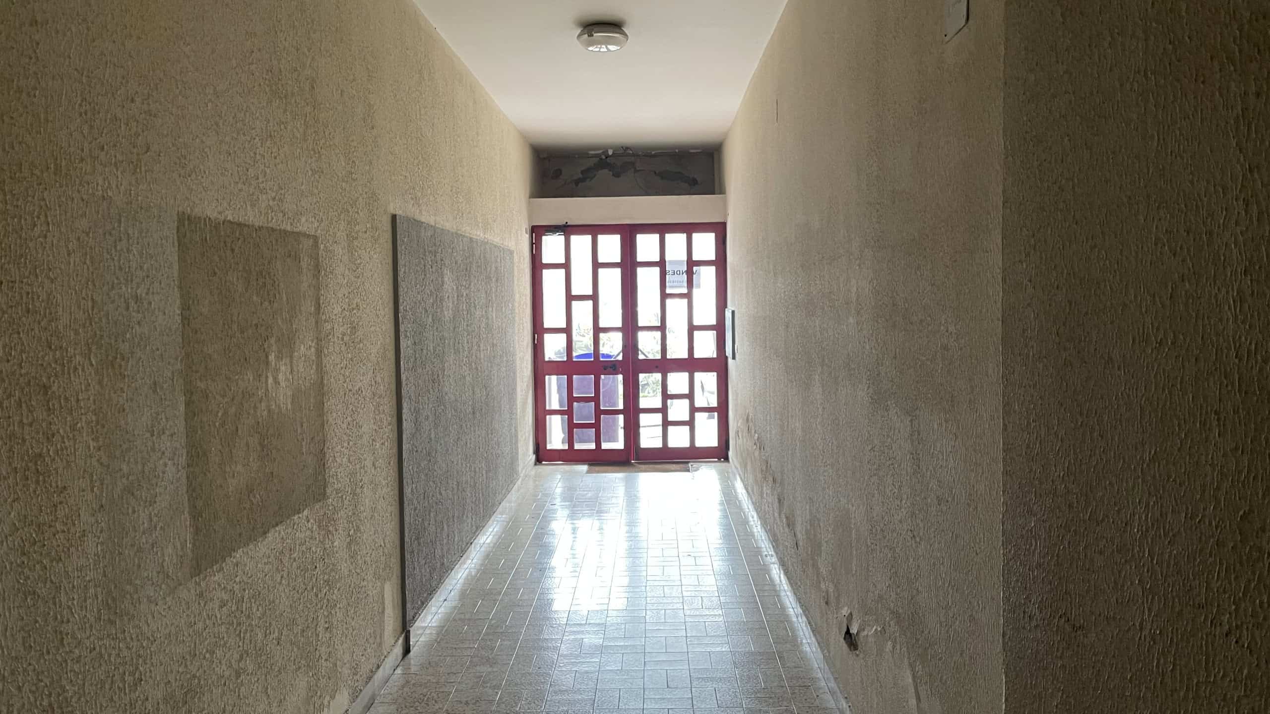 Ampio corridoio interno con pareti beige e pavimento in piastrelle lucide, illuminato dalla luce naturale proveniente da una finestra con grate rosse alla fine del passaggio.