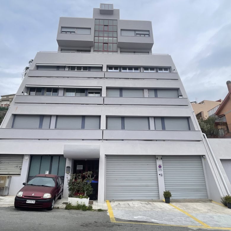 Immagine di un edificio residenziale moderno con più piani, situato in un'area urbana collinare, con veicoli parcheggiati davanti e un cielo nuvoloso sullo sfondo.