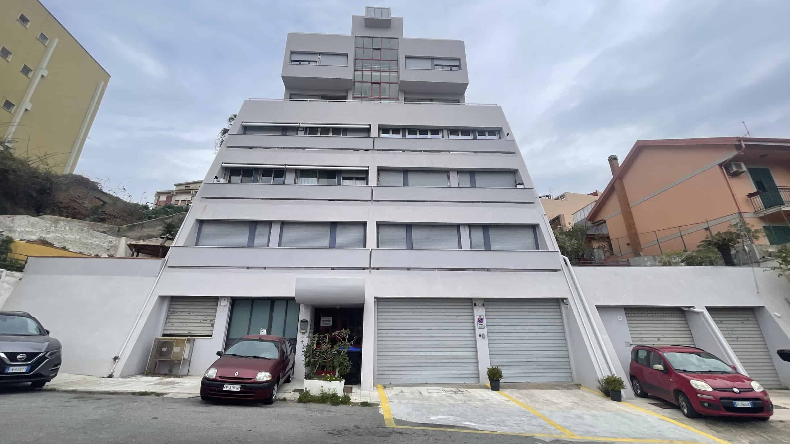 image00027 Immagine di un edificio residenziale moderno con più piani, situato in un'area urbana collinare, con veicoli parcheggiati davanti e un cielo nuvoloso sullo sfondo.