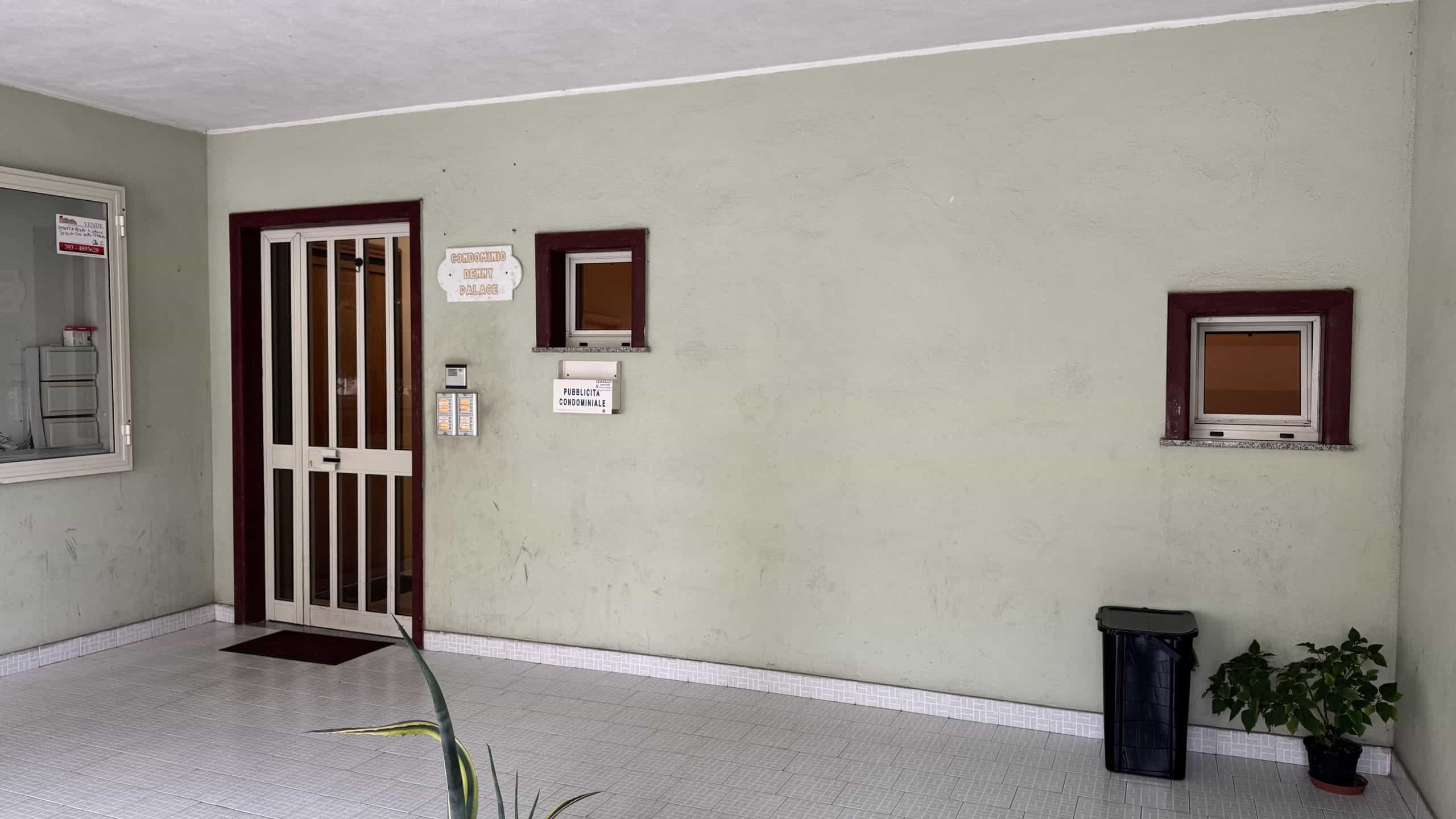 Cancello di sicurezza con portoncino in metallo e finestre piccole, ingresso condominiale con area di attesa e sistema di videosorveglianza, ambiente esterno pulito e ben curato.
