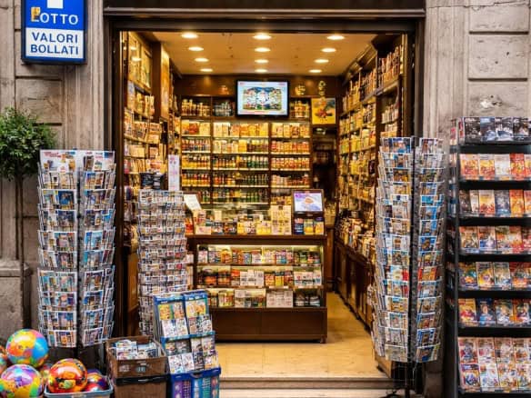 Negozio di tabacchi con vasta selezione di sigarette, cartine e articoli da fumatori, situato in una strada urbana con espositori di prodotti all'esterno.
